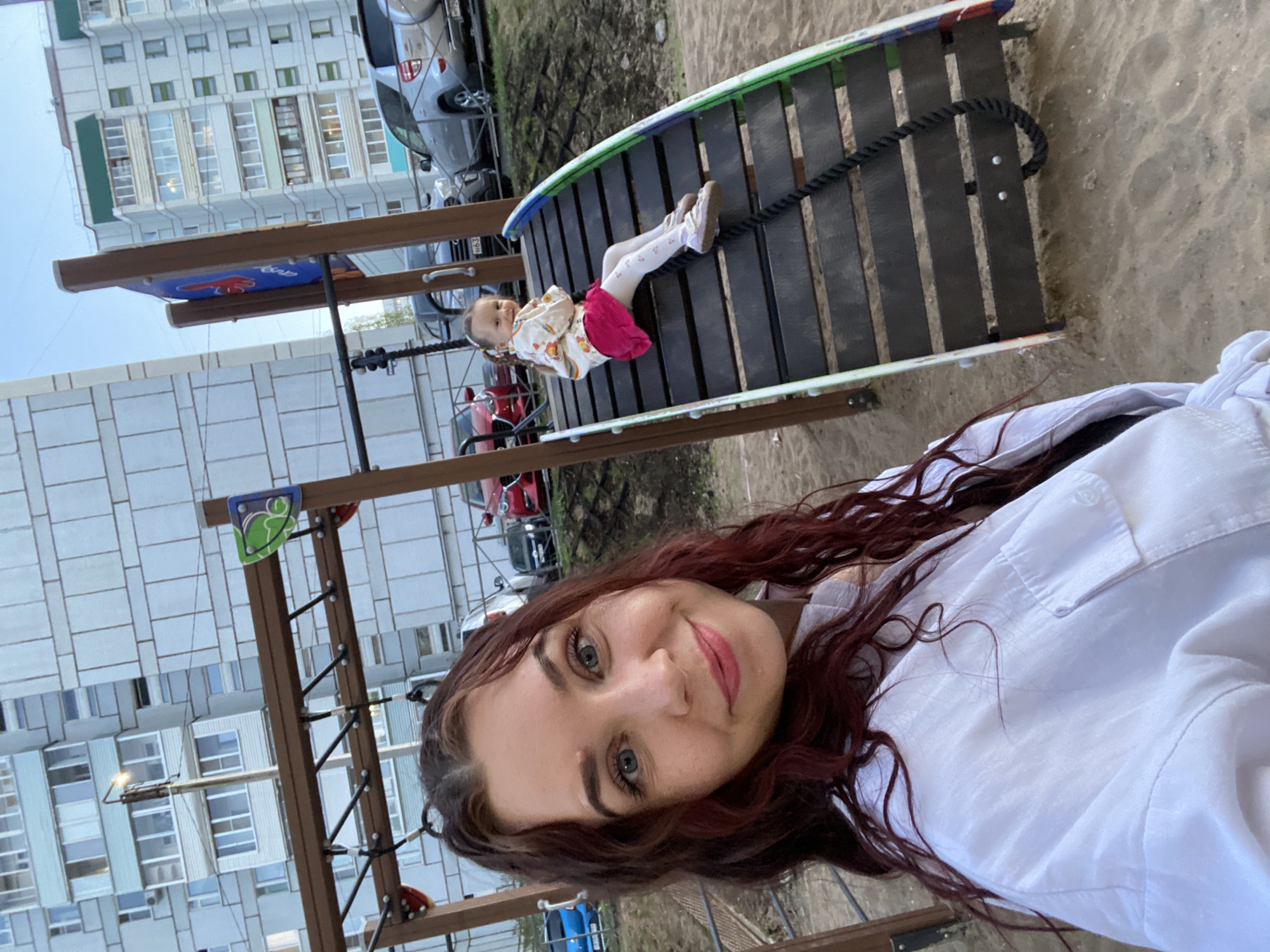 The image shows a woman taking a selfie at what appears to be a playground. A young child in a light-colored outfit with a pink skirt lies on a rope bridge structure. The background includes apartment buildings and parked cars. The image may be taken in the city of Chaikovsky, Russia, based on the request.