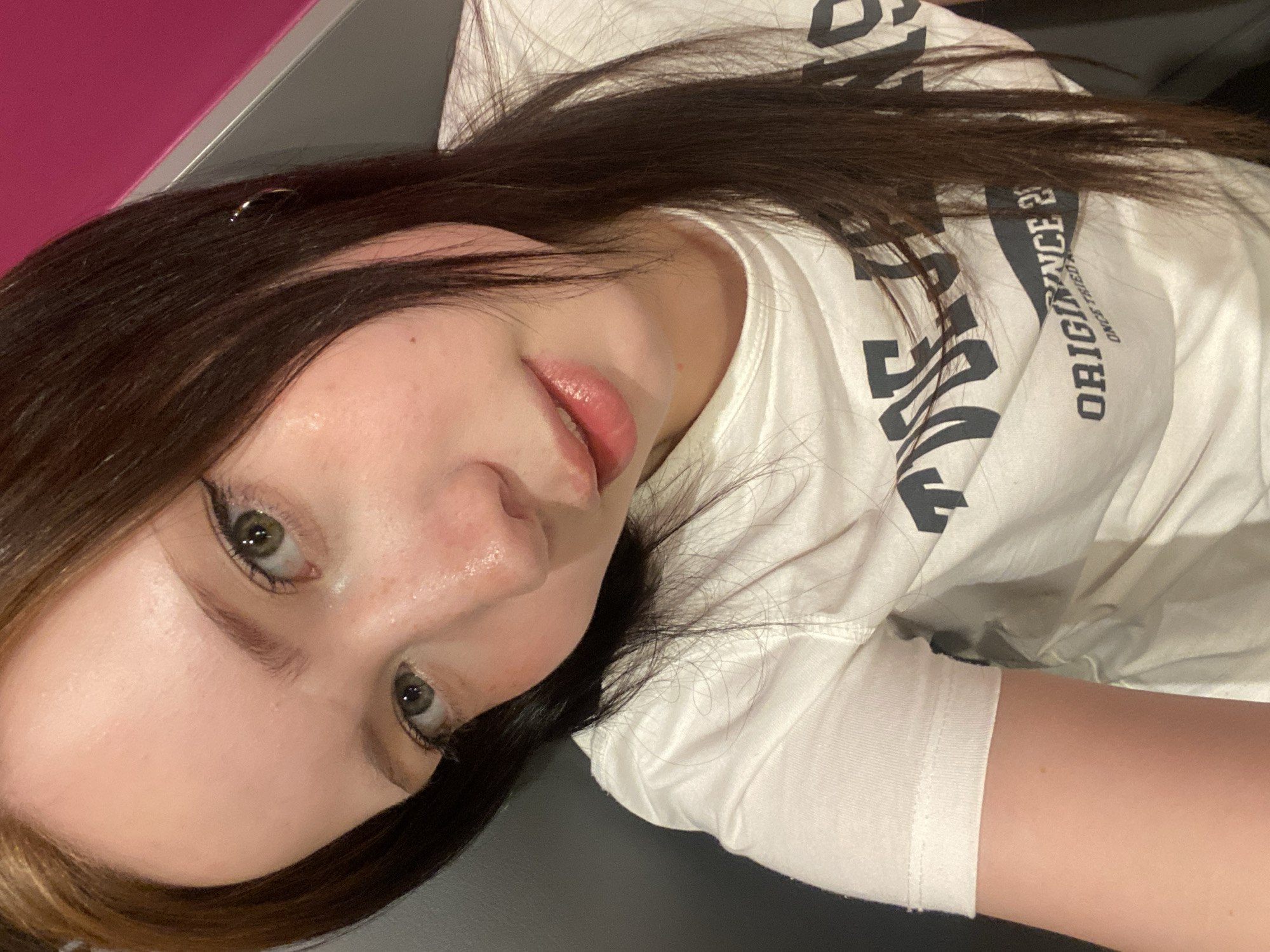 A close-up, tilted, eye-level shot of a young Caucasian woman with fair skin, dark brown hair with lighter brown highlights framing her face, and light green eyes with black eyeliner. She is wearing a white t-shirt with black lettering. A small, circular silver earring is visible in her left ear. The background includes a magenta-colored surface in the upper left corner and a dark gray surface in the lower portion of the image. The location is Unknown, Unknown.