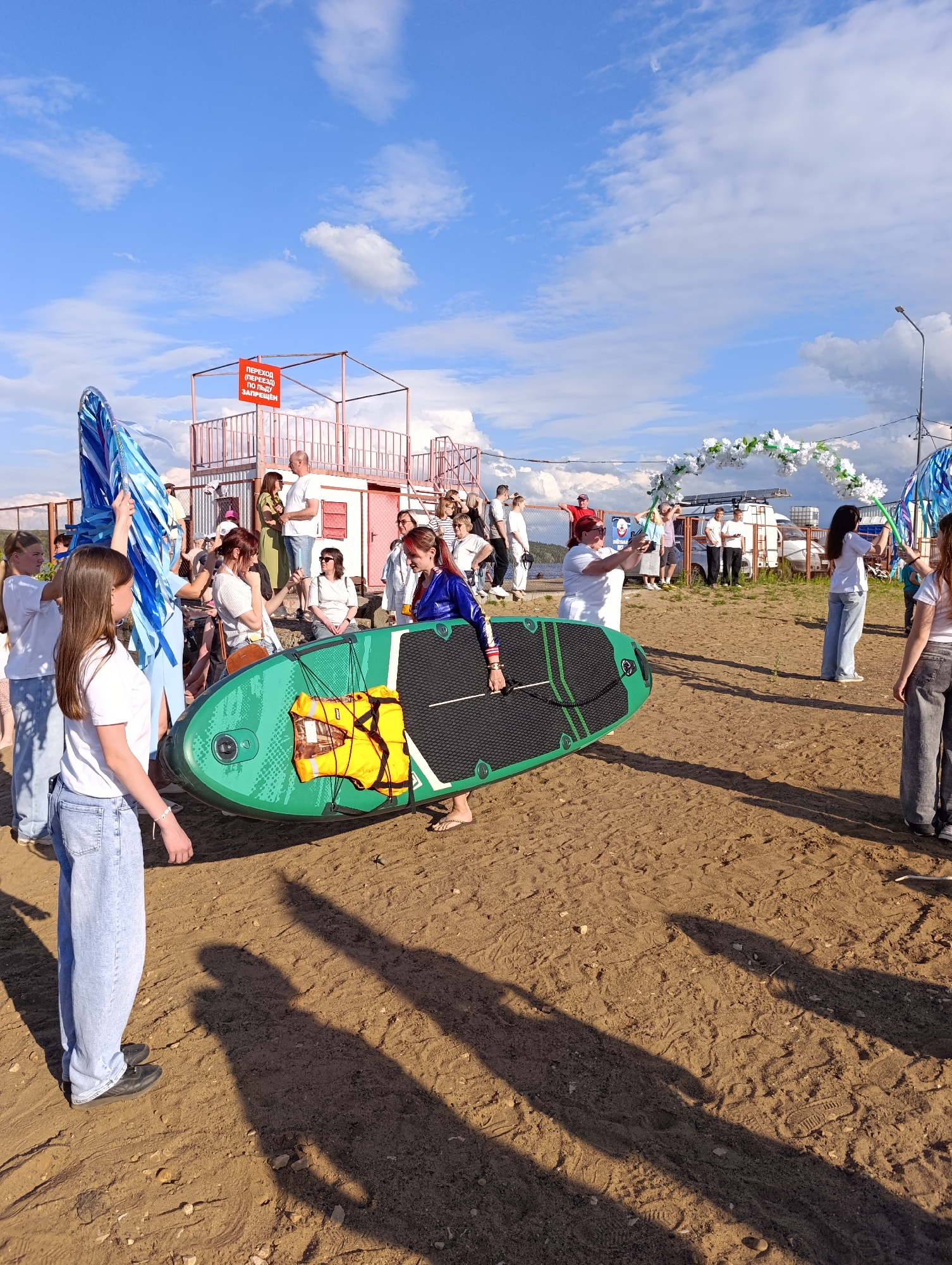 The image depicts an outdoor gathering on a sandy area, possibly a beach or riverbank, under a bright, partly cloudy sky. In the foreground, a young woman in a white t-shirt and light blue jeans is walking towards the left, carrying a large, green paddleboard with black accents. The paddleboard has an orange life vest attached. Behind her, several other people are visible, including a woman in a blue jacket and another in a white t-shirt, also looking towards the paddleboard. Further in the background, a group of people are gathered near a small, elevated structure with a red sign that has Russian text. To the right, a decorative arch adorned with white flowers is being held up by two people. A lamppost is visible in the far right background. The sandy ground is marked with footprints. The overall atmosphere appears to be casual and festive, possibly related to an event or celebration. The sign in the background reads "ПЕРЕХОД (ПЕРЕЕЗД) ПО ЛЬДУ ЗАПРЕЩЕН," which translates to "Crossing (moving) on ice is prohibited," suggesting this location is near a body of water that is frozen during winter months. This sign is characteristic of public safety regulations often found in Russian cities, and the context of a sandy area near water aligns with the geography of Chaikovsky, Russia, which is situated on the Kama River, a large river with a reservoir.