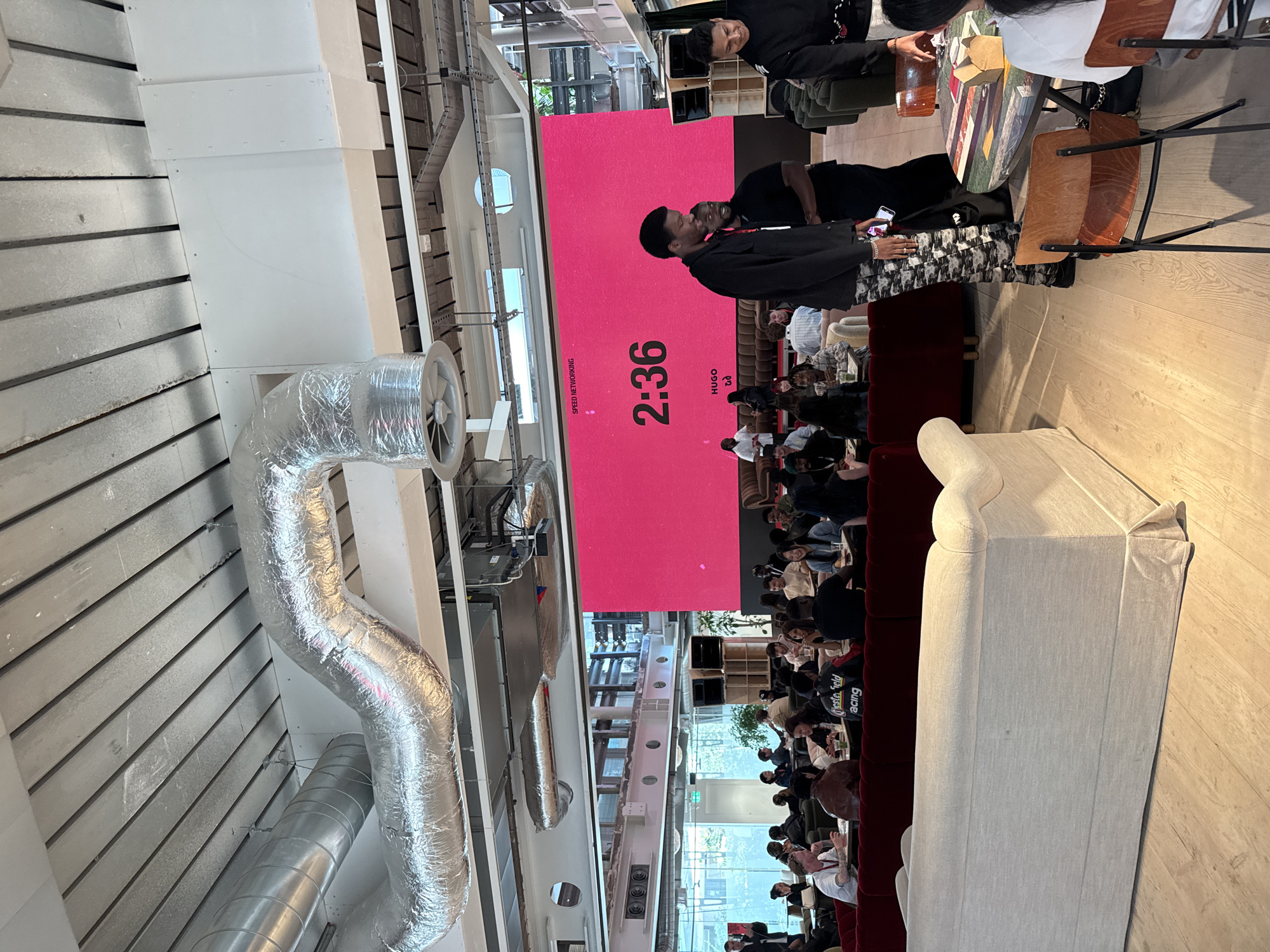 The image captures an indoor scene in a modern space, possibly in London, United Kingdom. A large screen dominates the scene, displaying a magenta color with the time "2:36" and the word "HUGO" underneath, likely indicating an event. Many people are seated in rows facing the screen, suggesting a presentation or conference. Some individuals are engaged in conversations at tables, with drinks and food items visible, hinting at a networking event. Architectural elements include exposed ductwork on the ceiling and large windows, giving the space a modern and industrial aesthetic. The image focuses on the interaction and the event, not on the exact location.
