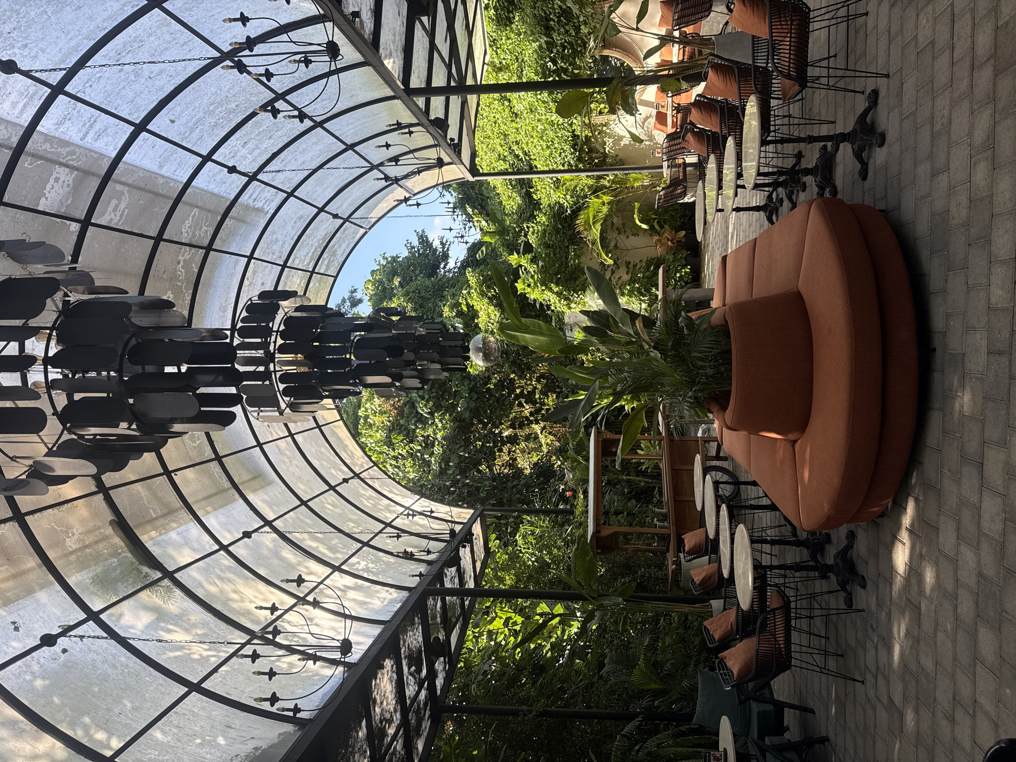 This image depicts the outdoor seating area of a restaurant or cafe with a covered, arched ceiling in Ubud District, Indonesia. The ceiling appears to be made of a translucent material stretched over a black metal frame, allowing dappled sunlight to filter through. Two large, modern chandeliers hang from the ceiling, along with several smaller, traditional-style candelabra chandeliers.
In the center of the seating area is a large, circular orange sofa with a planter in the middle, housing a tall green plant. Surrounding the sofa are several small, round marble-topped tables with black metal bases, each paired with black wire chairs with orange cushions. In the background, lush green foliage and trees are visible, suggesting a tropical environment. A wooden cart, possibly a bar or serving station, is also present. The floor is paved with gray rectangular tiles. The overall atmosphere is one of a stylish, semi-open-air dining or lounge space, integrated with abundant greenery. No people are visible in the image.