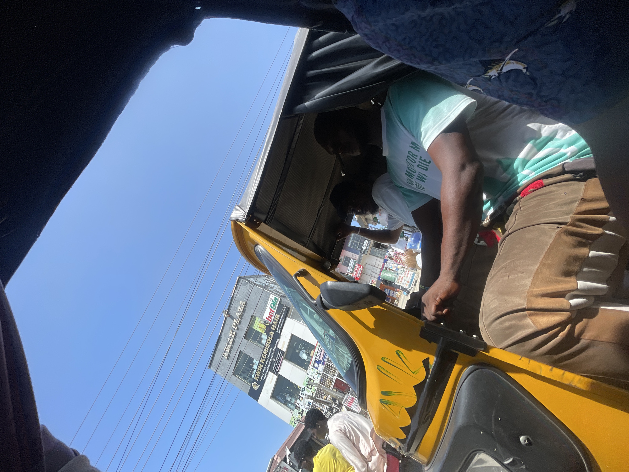 The image provides a candid, eye-level view from inside a yellow auto-rickshaw, commonly known as a Keke Napep, in Ado, Nigeria. The perspective is looking outwards, with the vehicle's dark fabric roof and side panels framing the upper and lower portions of the shot.
In the foreground, a person with dark skin is visible from the waist up, likely a passenger or the driver. They are wearing a light green t-shirt with white text that partially reads "MOTOR M YO WA DIE" and brown pants. Another individual is partially obscured behind them. The Keke Napep features yellow bodywork with black decorative paint.
The scene outside the vehicle is a vibrant urban environment bathed in bright sunlight under a clear blue sky. Numerous thin power lines stretch across the sky. Several multi-story buildings are visible in the background. A prominent grey building displays the large sign "AMAK PLAZA." Below it, a sign advertises "KWIN KEMISOLA HAIR," offering "Luxury Wigs & Hair Extension" and "Hair Installation Braiding Revamping," accompanied by a long numerical string (0010070101309012171аза). Other people and market-like stalls are discernible further down the street, suggesting a busy commercial area. The strong shadows indicate a sunny mid-day setting.