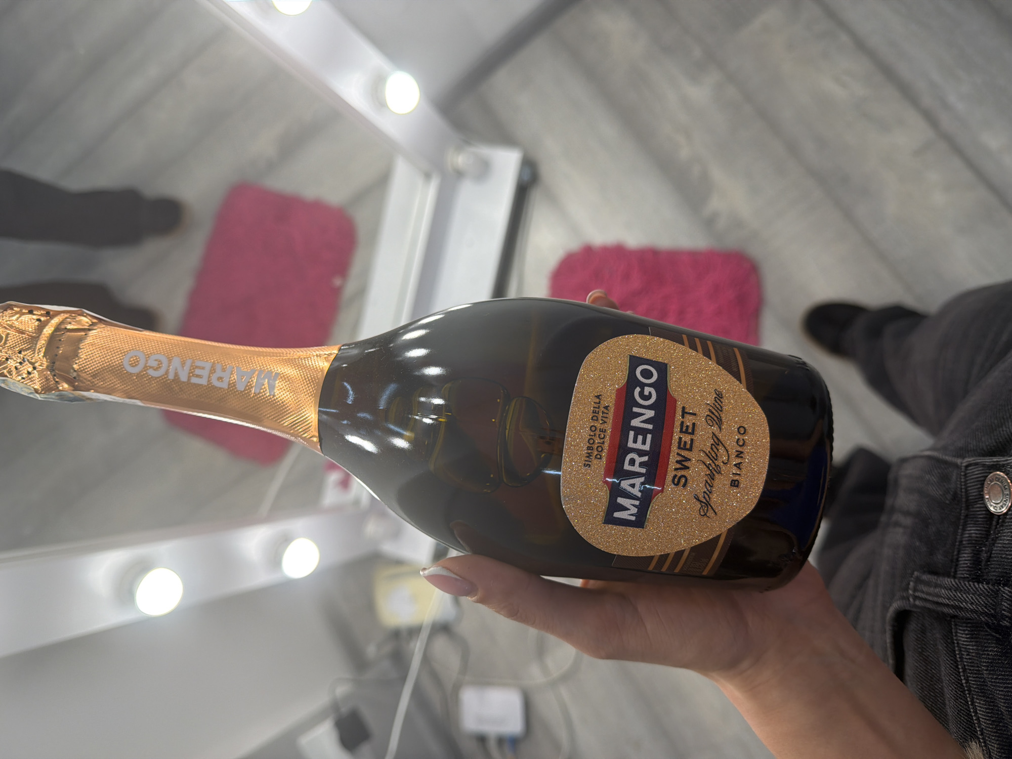 A person's hand holds a bottle of Marengo Sweet Sparkling Wine Bianco, with a gold foil cap and label adorned with glitter. The background includes a brightly lit mirror with lights, a pink fluffy rug, and a floor that appears to be wooden planks. The setting is indoors, likely a personal space, and does not provide any specific indicators of a location within the city of Kyiv, Ukraine.