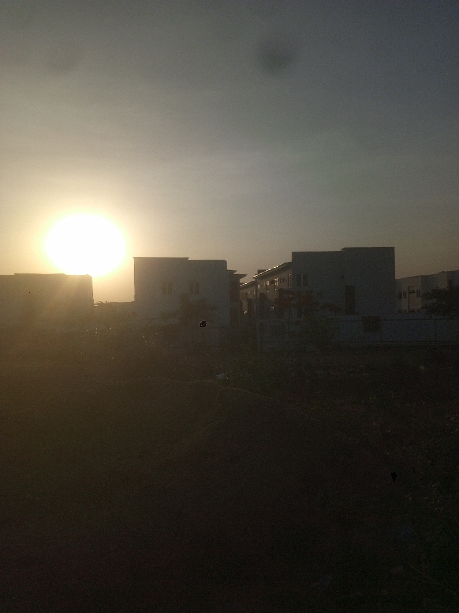 The image shows a cityscape at sunset in Gwarinpa, Nigeria. In the foreground, the ground is dark and obscured, possibly vegetation or a pile of earth. Several white or light-colored buildings are visible in the midground, with a modern, multi-story architectural style. The sun dominates the sky, appearing as a bright orb with a hazy glow, positioned to the left of the buildings. The sky fades from a lighter shade near the sun to a darker hue toward the top of the frame. There is no visible evidence of people, actions, or interactions in the frame.