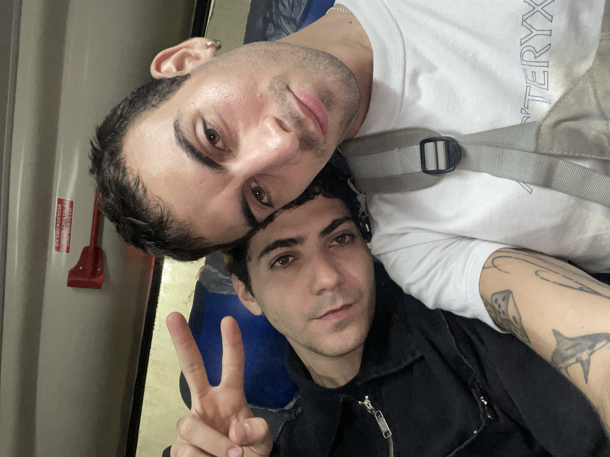Two young men are posing for a selfie. One man wears a white t-shirt with the word "ARCTERYX" printed on it and has a grey backpack strap over his shoulder. He has a hoop earring in his left ear. The other man wears a black jacket and is holding up a peace sign. The photograph appears to be taken inside a train or bus, given the emergency window breaking tool visible on the left side of the frame. The location is unknown, and there is nothing visible in the image that definitively places it in Buenos Aires, Argentina.