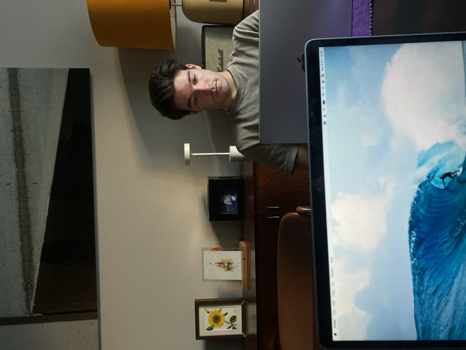 This image depicts an indoor scene, likely a home or office, situated in Lordelo do Ouro e Massarelos, Portugal. In the foreground, the top half of a laptop screen is visible, displaying a wallpaper of a surfer riding a large blue wave under a partly cloudy sky, with the macOS menu bar showing "Mon 6 Oct 18:05". Behind this, a young adult male with dark hair, wearing a light-colored t-shirt, is seated and intensely focused on a silver laptop positioned directly in front of him. A purple fabric mat is visible beneath his laptop. In the background, a dark wooden cabinet/sideboard holds several items: a framed picture of a sunflower, another framed artwork, a small black digital photo frame, and a white minimalist table lamp. To the right, an orange-shaded table lamp with a brass-colored base is present, and a "Marshall" branded amplifier or speaker is partially visible behind the male's head. The wall behind the cabinet is light grey, with a large abstract art piece featuring geometric shapes in dark grey, black, and light grey mounted higher on the wall. Part of a brown chair is visible on the left side of the frame.