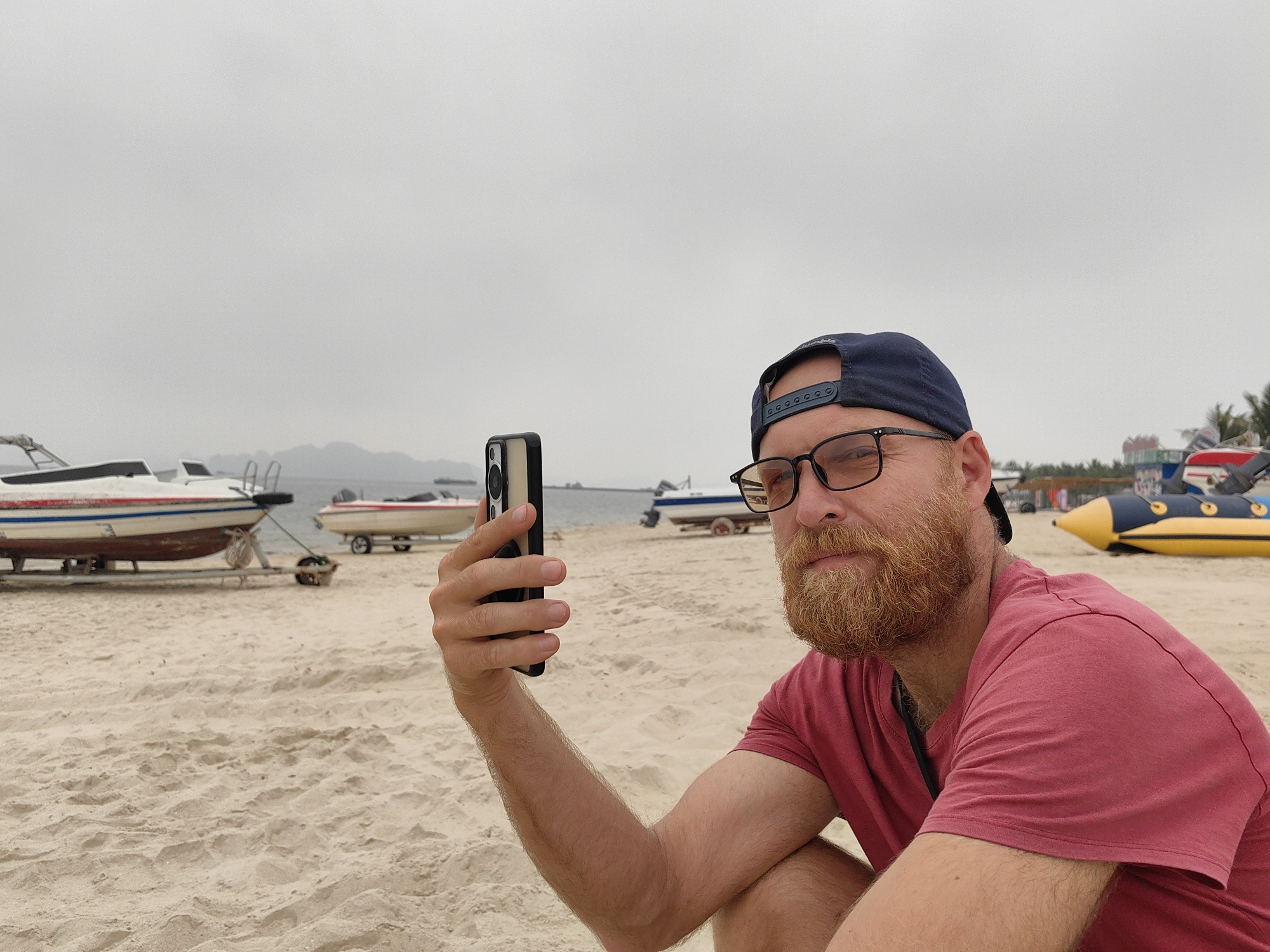 A man is on a sandy beach in Bai Chay Ward, Vietnam, holding up a smartphone. He is wearing a red t-shirt, glasses, and a navy blue baseball cap, and has a reddish beard. In the background, there are several boats on the beach, and the sky is overcast. A yellow and black inflatable banana boat is to his right.
