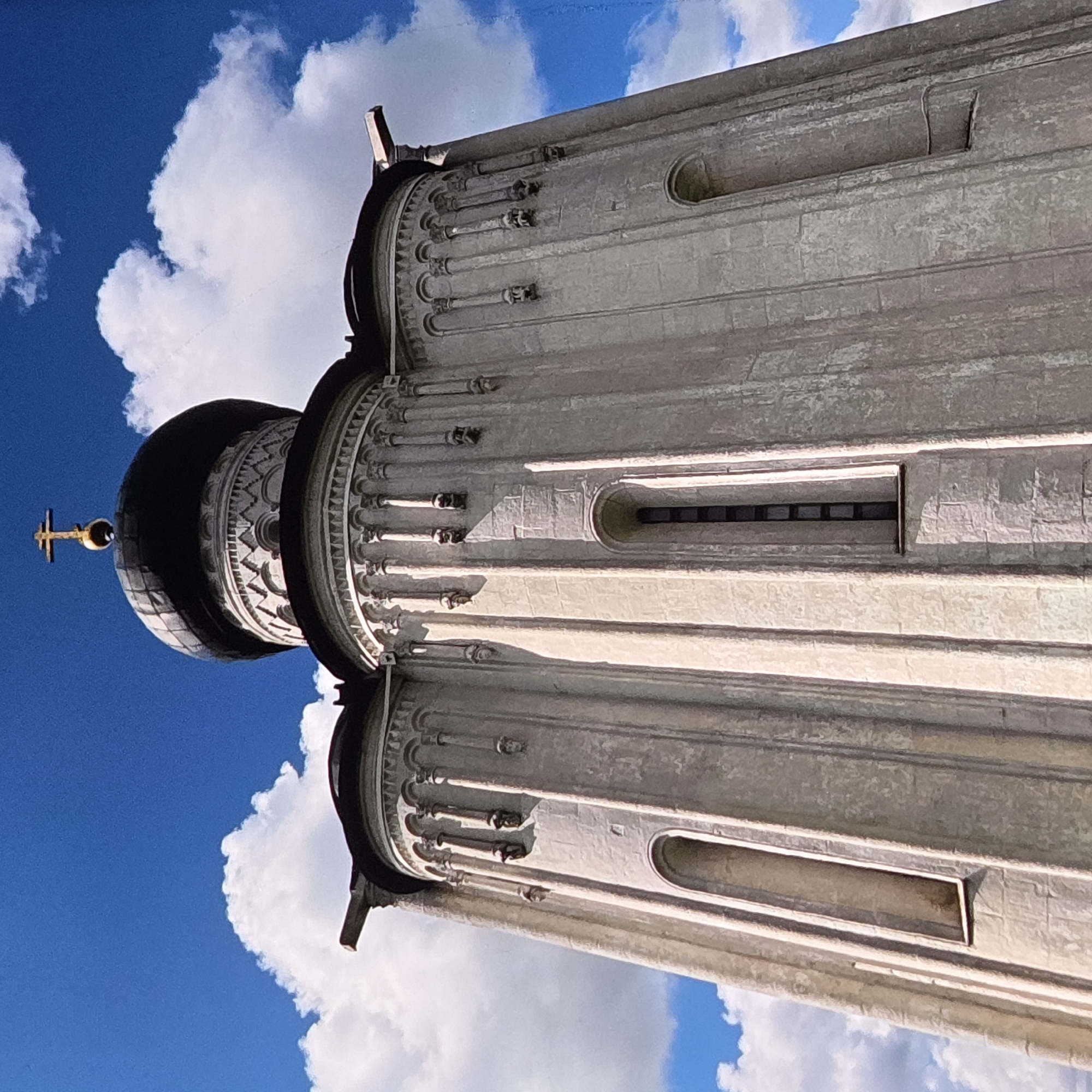 The image shows the Church of the Intercession on the Nerl, located in Vladimir, Russia. The church, a UNESCO World Heritage Site, is constructed of white stone and features a single onion dome topped with a golden cross. The building has a distinct architectural style with multiple semi-circular apses and decorative blind arcades. Several small, carved faces or gargoyles are visible on the exterior walls. The church is set against a bright blue sky filled with fluffy white clouds. The low-angle shot emphasizes the grandeur and height of the church.