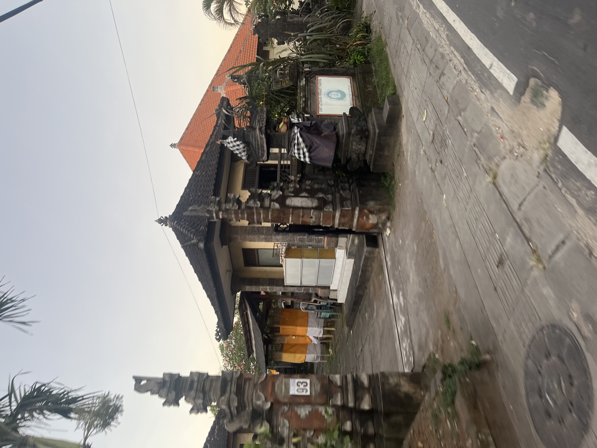This image captures a street view in Desa Adat Kutuh, Kuta Selatan, Bali, Indonesia. The scene is illuminated by natural light, suggesting either late afternoon or early morning, with a pale blue sky transitioning to a light yellowish-orange tint at the horizon.
In the foreground, a paved sidewalk with tactile strips and a circular metal manhole cover is visible on the left. The sidewalk is bordered by a concrete curb and an asphalt road, marked by a solid white line.
The central feature is the entrance to a property, framed by two ornate, dark stone gate pillars characteristic of traditional Balinese architecture. The left pillar displays the white numerals "93." Beyond the gate, a paved path leads into the property. To the left, a sheltered structure contains two upright objects draped in yellow and white fabric. A building with a dark tiled roof is partially visible behind these drapes, and another, lighter-colored building with a dark tiled roof is present further back on the right.
To the right of the property entrance, a stone shrine (pelinggih) is prominent, adorned with a black and white checkered fabric (poleng). Adjacent to this, a white sign with blue text and a circular logo is mounted on a low stone wall. The text on the sign reads "UNIT USAHA PIRANTI YADNYA" and "DESA ADAT KUTUH." The property perimeter is lined with various green plants and bushes, including taller leafy vegetation near the sign. In the background, a building with a distinctive orange tiled roof is visible. Palm fronds extend into the upper left quadrant of the frame. No individuals are visible in the scene.