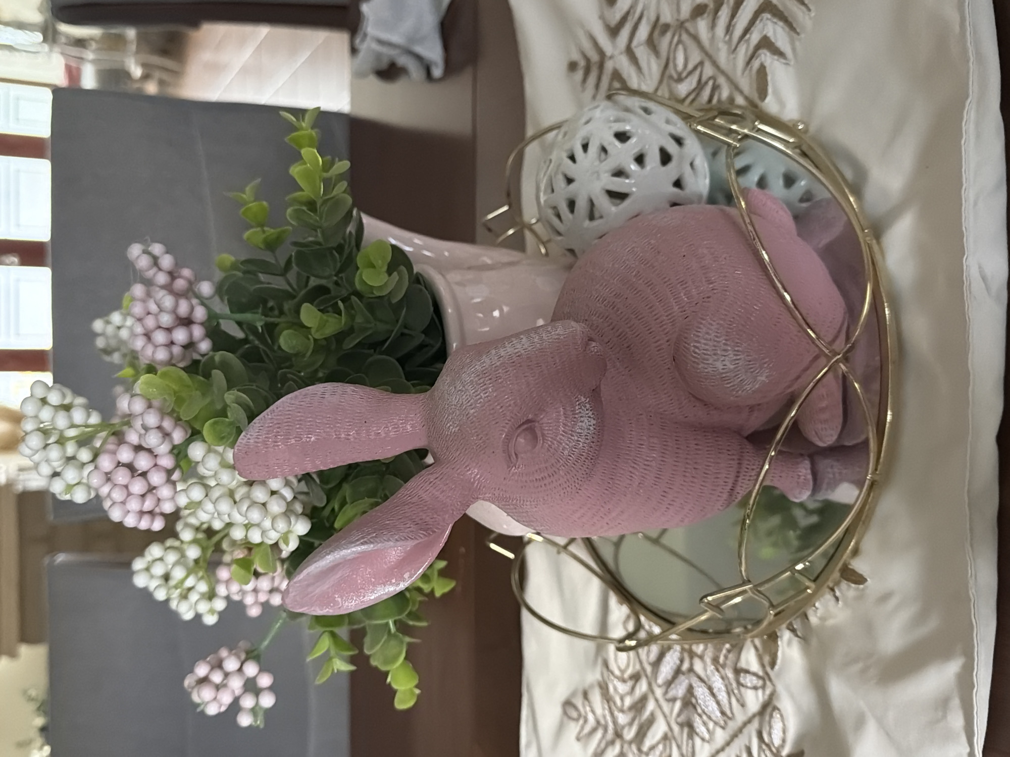 A close-up shot of a decorative Easter arrangement. The centerpiece includes a pink bunny figurine in a gold metal frame, placed on a white tablecloth with an embroidered pattern. A white vase filled with green foliage and white-and-pink berry-like decorations sits next to the bunny. A white decorative ball with cut-out patterns is also visible. The scene appears to be indoors. There is no indication of the location being Cuautla, Mexico.