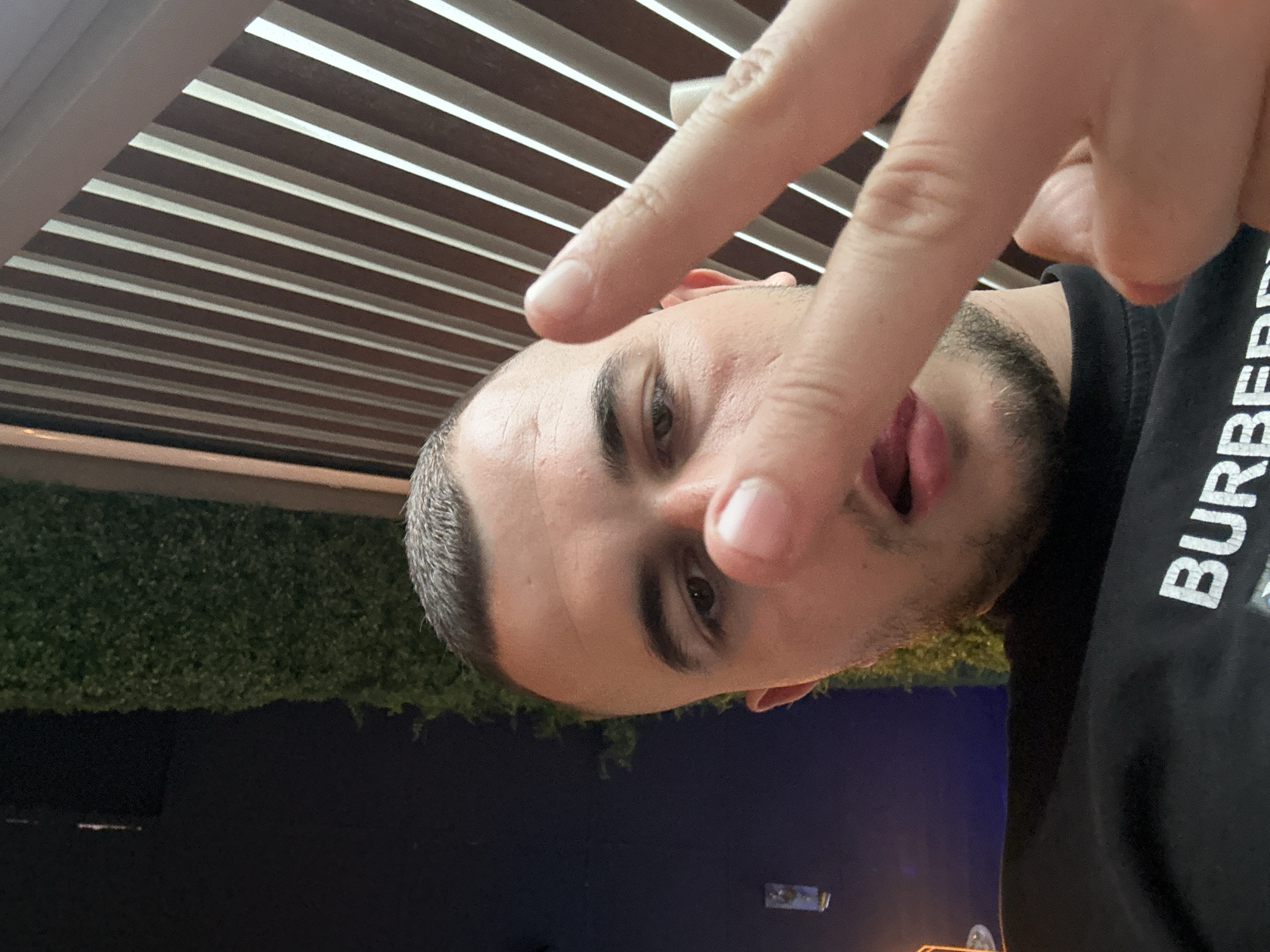 A man is taking a selfie. He has short dark hair, a dark beard, and is wearing a black t-shirt with "BURBERRY" printed on it. He is making a peace sign with his fingers close to the camera. The background includes a dark area possibly reflecting outdoor lights, some greenery, and a striped ceiling. The location is Plovdiv, Bulgaria.