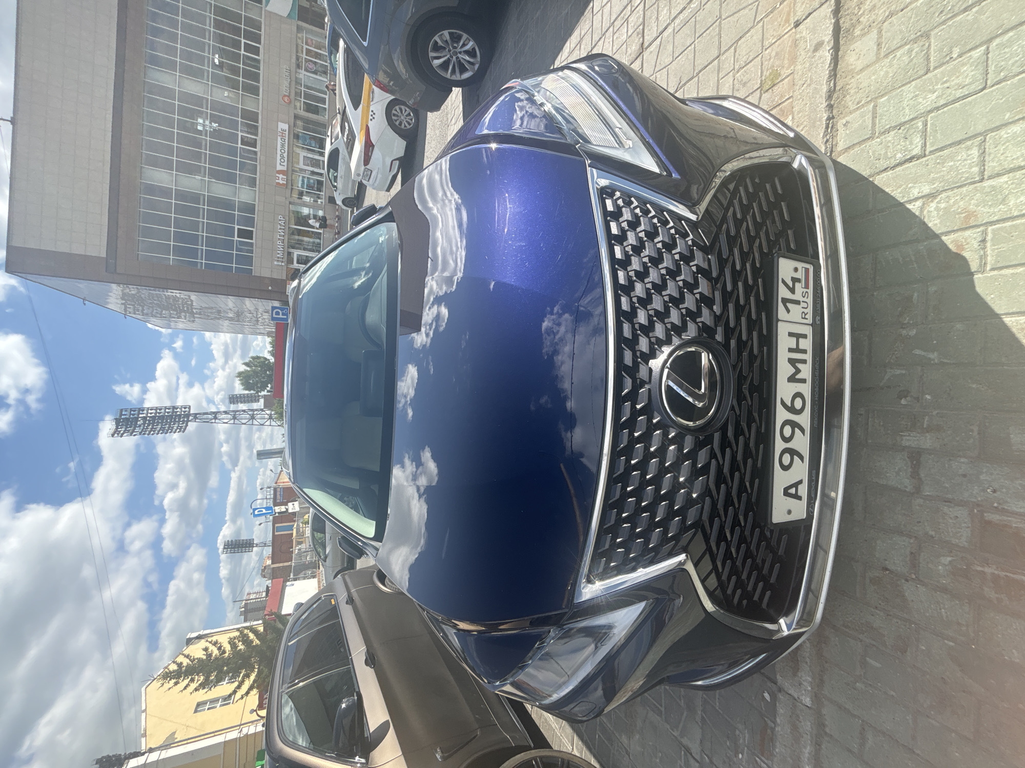 In a street-level, full-frame, eye-level shot, a dark blue Lexus RX SUV is parked on a paved surface. The SUV's front grille, headlights, and license plate are prominently visible. The license plate reads "A 996 MH 14" and "RUS." The vehicle is parked in what appears to be a commercial or public area, indicated by a large, modern building with glass windows in the background. Signs on the building include "МИР ГИТАР" (World of Guitars), "ГОРОЖАНКА" (Gorozhanka), and "КОЛПИЦЕНТР" (Kolpcenter), suggesting a shopping center or complex. Several other vehicles, including a white taxi with a yellow stripe, are also parked in the vicinity. The sky above is blue with scattered white clouds, and tall light towers, possibly from a stadium, are visible in the distance. The scene is lit by sunlight, casting shadows on the ground.