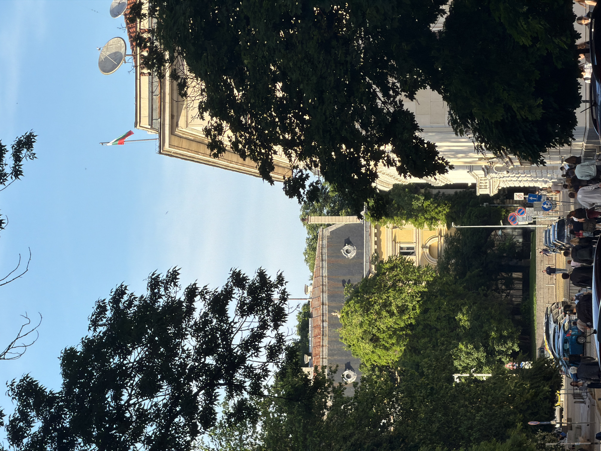 The image captures a vibrant street scene in Sofia, Bulgaria, characterized by historic architecture and lush greenery under a clear blue sky. The perspective looks slightly upwards and across a street, revealing the upper portions of buildings and the sky above, partially obscured by trees.
On the left side of the scene, a grand, light-colored building with ornate classical details is prominent. From its corner, the national flag of Bulgaria, featuring horizontal stripes of white, green, and red, proudly flies. On the roof of this building, two large satellite dishes are visible.
In the foreground and mid-ground, dense green foliage from large trees frames the view, with sunlight dappling through the leaves. Across what appears to be a street, another distinct building is visible through the trees, featuring a dark, sloping roof with several circular dormer windows. This building appears to be yellow or light-colored.
On the right side of the image, a bustling street and sidewalk are partially seen. Several people are walking or standing on the pavement. A line of parked cars is visible along the curb. Various street signs are present, including a red-bordered "No Parking" sign, a blue directional arrow, and a circular speed limit sign displaying "30".
The scene suggests a sunny, pleasant day, likely late morning or afternoon, given the bright illumination and the shadows cast. The clear sky and full green leaves indicate warm weather, probably spring or summer. The overall atmosphere is that of a typical, active city street.