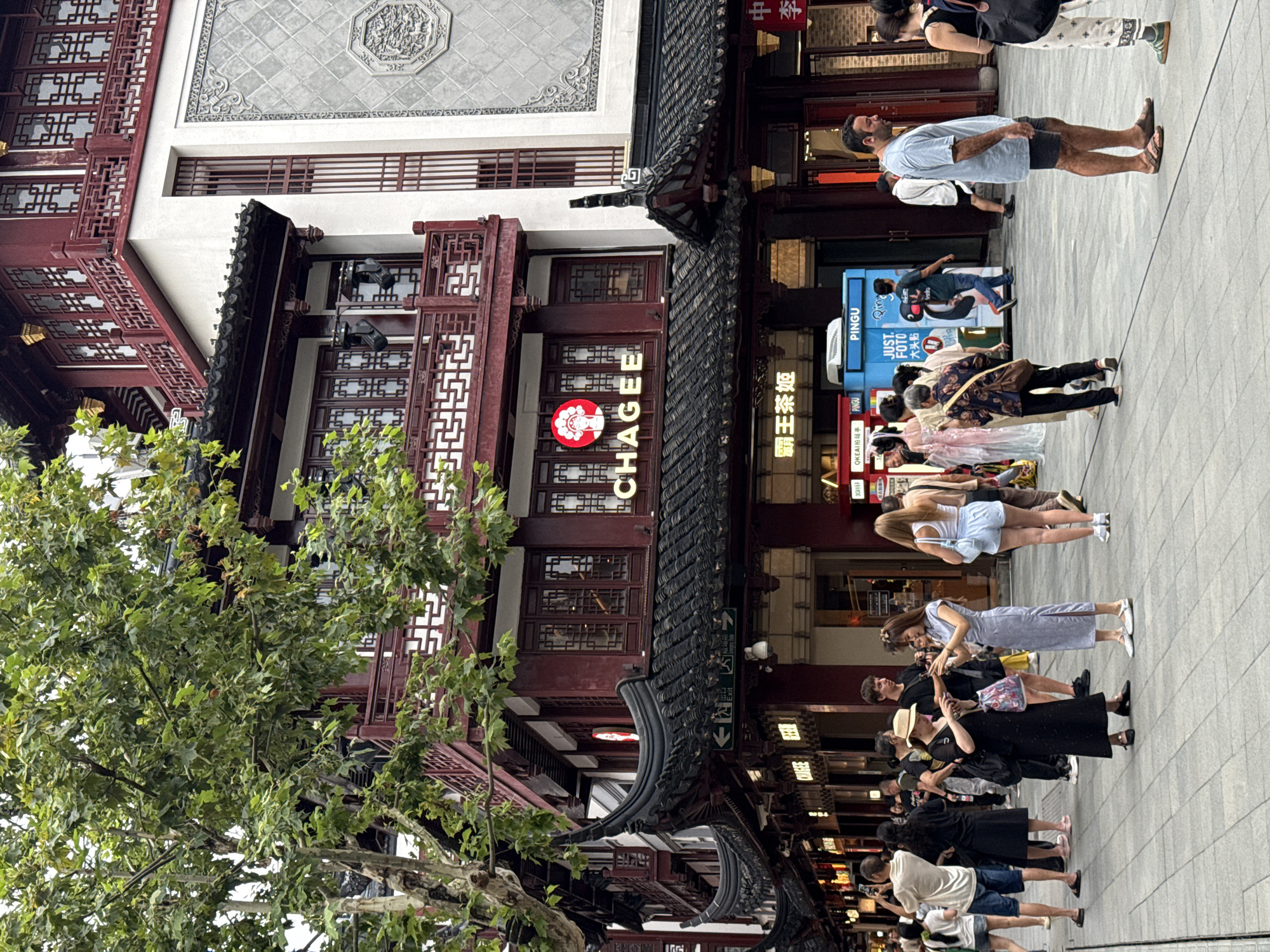 The image depicts a bustling street scene in Shanghai, China, likely within the Yu Garden Bazaar area, characterized by traditional Chinese architecture and a lively crowd. In the foreground, several individuals are seen walking and interacting. To the left, a group of people are gathered, some taking photos. A man in a light blue t-shirt and dark shorts is standing in the middle ground, facing right and engaged in conversation with a woman wearing a black top and patterned pants. Behind them, a blue kiosk with the "PINGU" logo is visible.
The architecture dominating the background features multi-story buildings with distinctive red facades, dark tiled roofs with upturned eaves, and intricate wooden latticework on balconies and windows. A prominent sign with the name "CHAGEE" in English and Chinese characters, along with a circular logo featuring a stylized face, is affixed to one of the buildings. Trees with lush green foliage overhang the street, providing shade. The pavement is made of light-colored rectangular tiles. The overall atmosphere suggests a popular tourist destination or commercial area within Shanghai, blending traditional aesthetics with modern commerce.