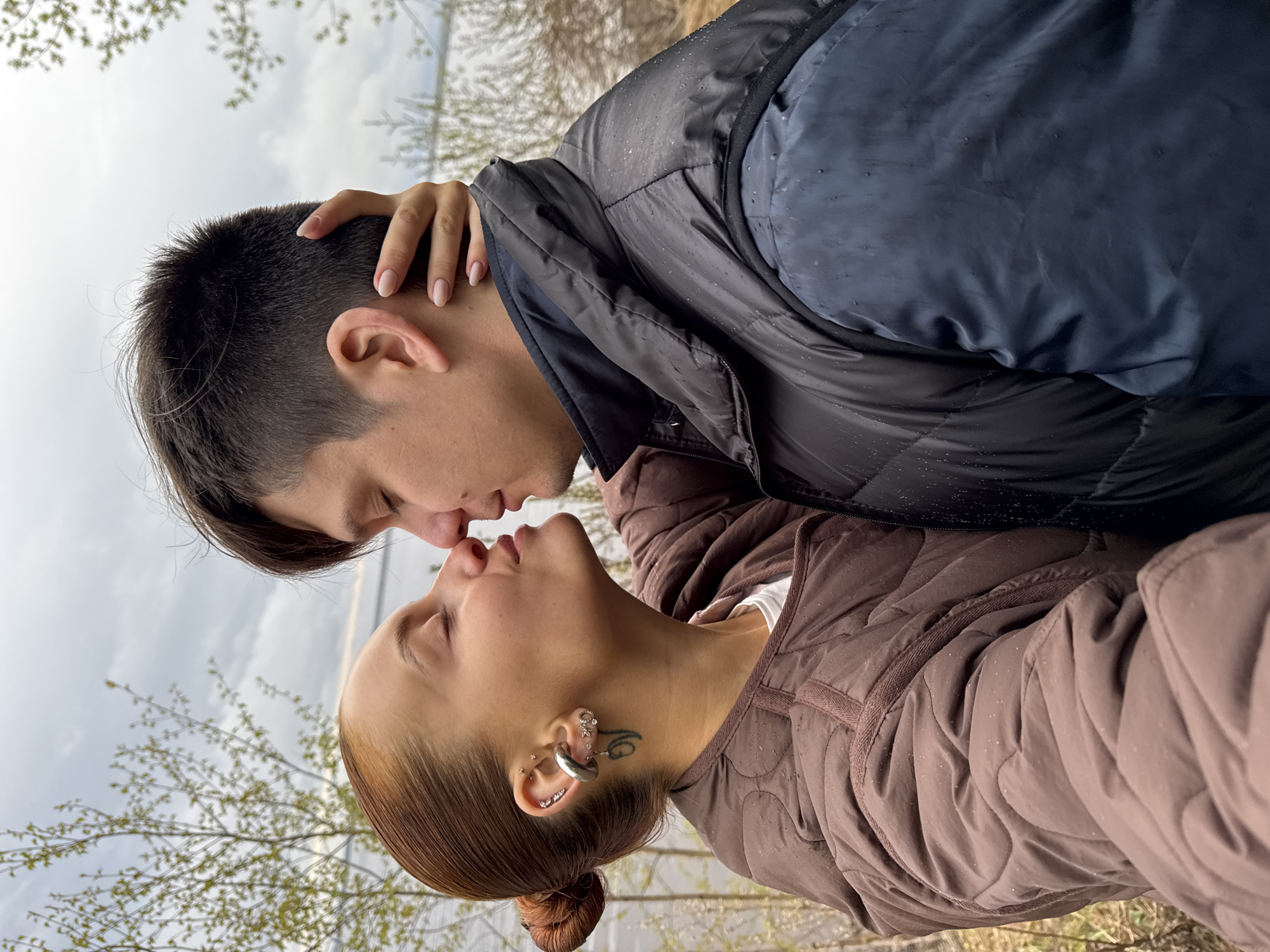 A couple stands in Chastyye, Russia, their faces close, appearing to kiss. The man, with dark hair styled with a side part, wears a dark blue and black puffer jacket. His hand rests on the back of the woman’s head. The woman, with reddish-brown hair in a bun, wears a brown quilted jacket and multiple piercings in her left ear, as well as a tattoo behind her ear. The background shows a blurred landscape with trees and a body of water under a cloudy sky. Raindrops are visible on the jackets.