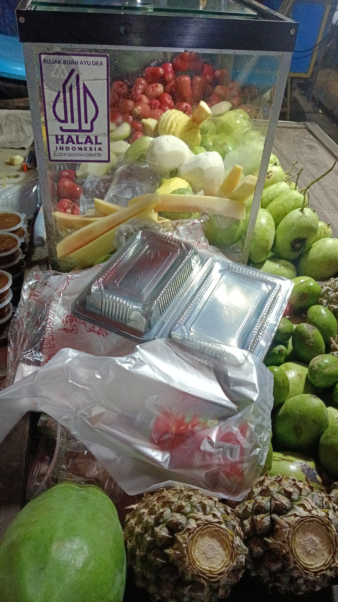 A close-up, medium shot captures a fruit stand in Tajurhalang, Indonesia, featuring a glass display case filled with a variety of fresh fruits. The display case is adorned with a purple and white "HALAL" sticker with Indonesian text, indicating the food's certification. Inside the case, chopped guava, rose apples, and possibly mangoes are visible, some appearing peeled and cut into segments. Alongside the display are several clear plastic containers, some stacked, and a plastic bag containing more fruit. In the foreground, two pineapples with their spiky exteriors and cut tops are prominently displayed, along with a whole green papaya. The scene is likely outdoors, given the lighting and the informal setup typical of street vendors. The overall impression is one of a vibrant, fresh fruit offering ready for sale.