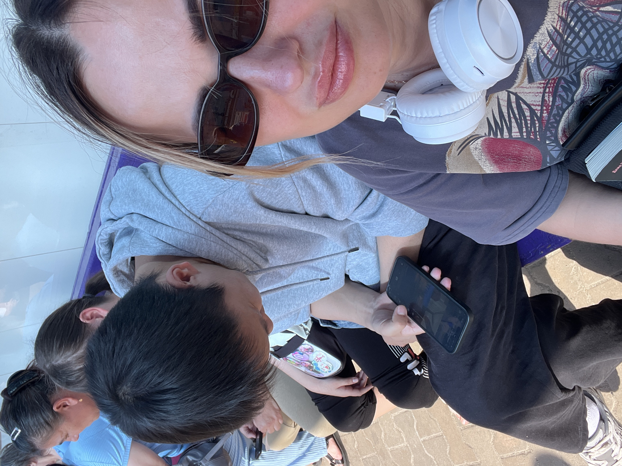 In an outdoor setting with a paved ground and a tiled wall in the background, several individuals are seated on a purple bench. The closest individual, a woman with blonde hair and sunglasses, is looking forward, her face partially visible. She is wearing a dark t-shirt with a graphic print and has white headphones around her neck. Next to her, a person with dark hair and a grey hoodie is looking down at a smartphone held in their hand. To their left, another person with dark hair is also looking down, their hand holding a smartphone. Further back, a woman in a blue shirt is visible, and another person's arm and leg are visible. The scene appears to be a public space, possibly a waiting area or a park bench.