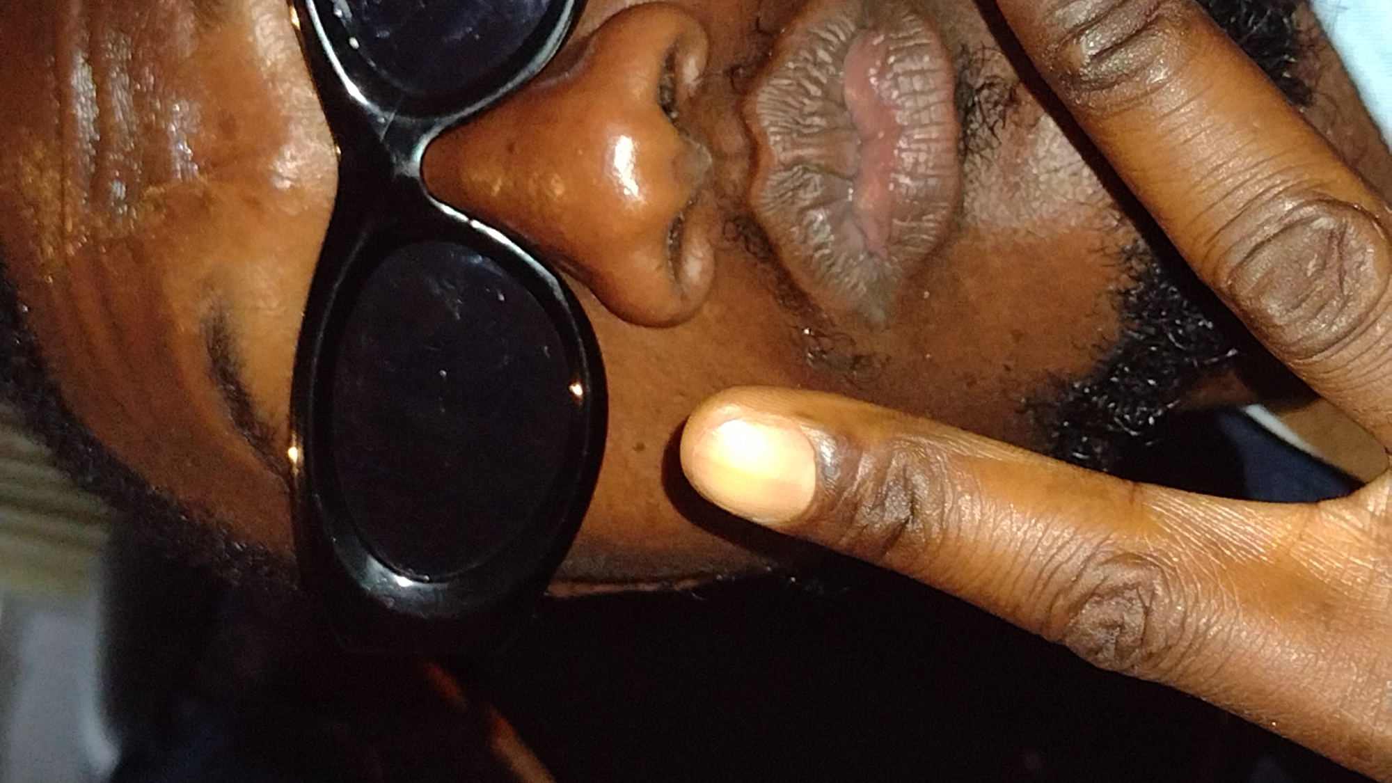 The image is a close-up, angled shot of a person's face, partially obscured by sunglasses. The person's skin tone is dark. They are wearing large, oval-shaped black sunglasses. The lower half of the face is visible, showing full lips pursed outwards. A finger is held up near the mouth, with the nail visible. The background is dark and out of focus. There is no indication of the city of Jos, Nigeria, or any specific environmental details that would place the image there. The image appears to be a selfie taken indoors or in a dimly lit area.