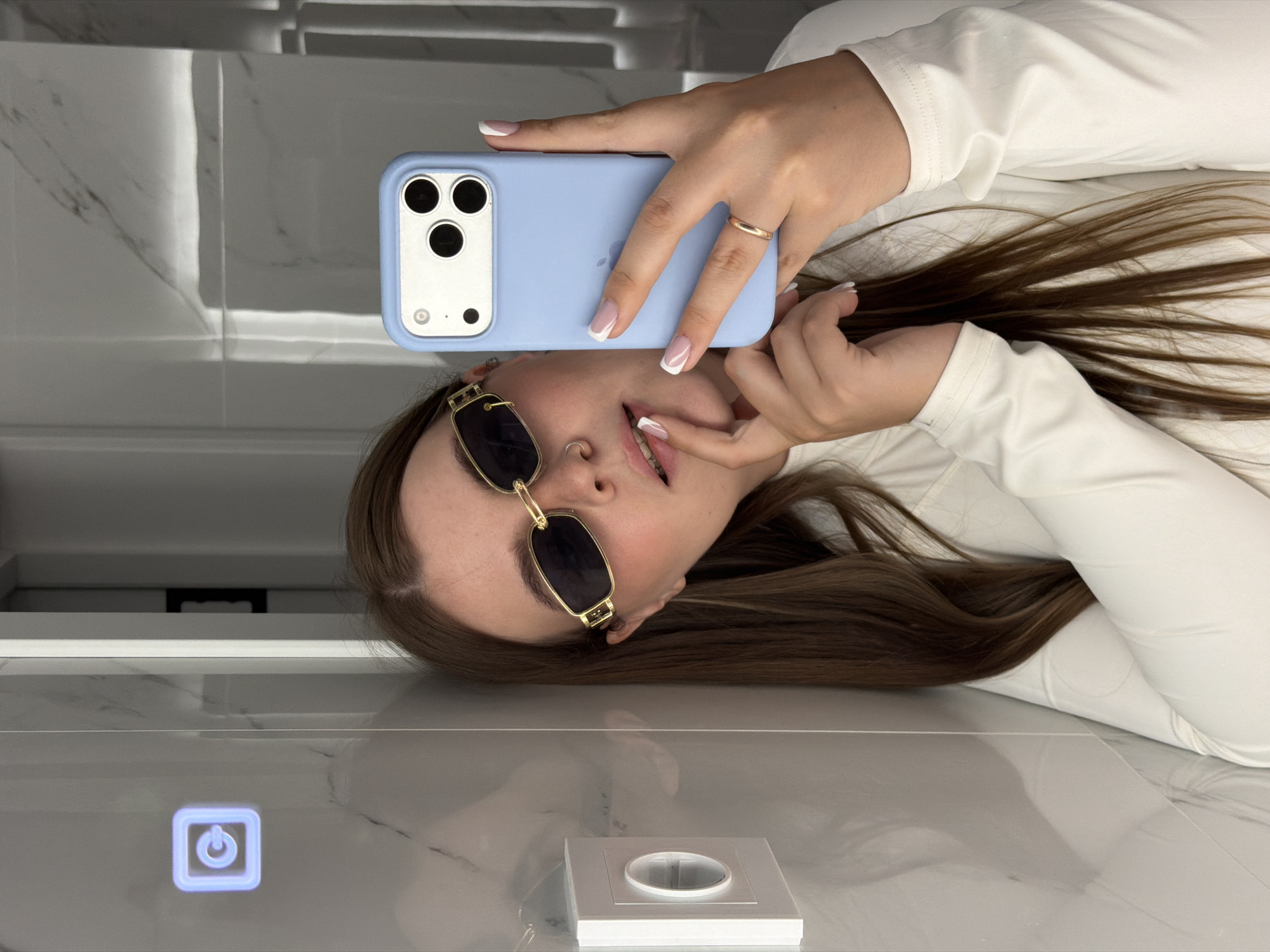 A woman is taking a selfie in a bathroom. She's wearing sunglasses and a white top, and she is holding a light blue smartphone in her right hand. The reflection of the woman and her phone can be seen in the bathroom mirror. The bathroom has a white and grey color scheme with marble tiling. A power button is illuminated on the mirror, and a light switch is on the counter. The image was likely taken indoors in the city of Chaikovsky, Russia.