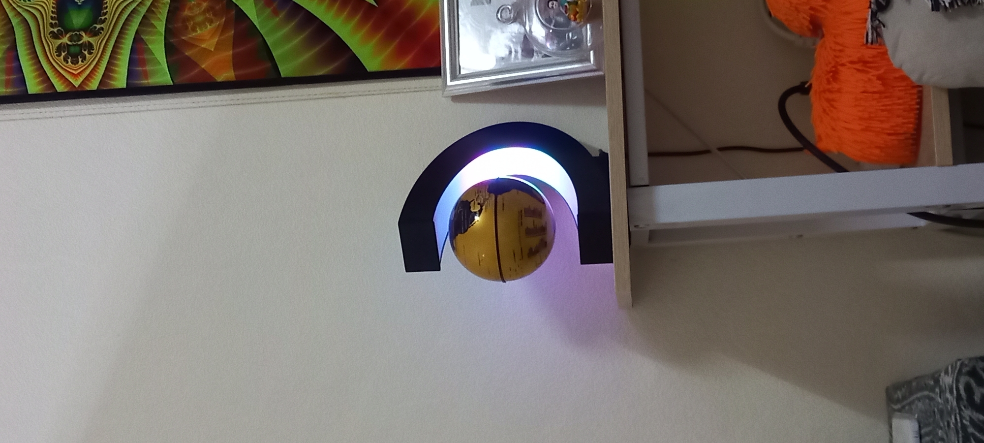 The image displays an indoor decorative arrangement, likely within a room in Baan Chaloklum, Thailand. A central feature is a levitating golden globe, patterned with black outlines resembling continents, suspended within a black arched stand. The stand emits a soft, gradient LED light, transitioning from white at the top to blue and purple towards the base, casting a purple glow and shadow on the wall.
Above this display, a framed abstract artwork hangs on the wall. The artwork features vibrant fractal-like patterns in shades of green, orange, red, and yellow. To the right, a light-colored wooden shelf extends into the frame. On the shelf, several items are visible: a small, silver-framed clear object, possibly a clock or display piece, and a large, fluffy orange object, likely a stuffed animal or decorative pillow, partially obscuring black cables. A portion of a white and grey stuffed animal is also visible on the shelf. The wall behind these items is a consistent light color, possibly white or light beige. A small section of a dark, patterned object, possibly furniture or a cushion, is visible in the bottom right corner. No visible text or explicit activity is present. The artificial lighting from the globe suggests the scene could be viewed at any time of day.