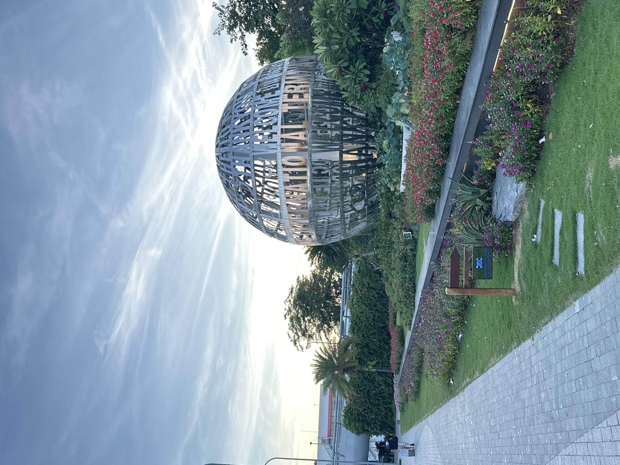 The image shows a large, spherical sculpture made of metal latticework. The sculpture is covered in what appears to be text, but it is difficult to read clearly. It is situated in an outdoor setting with lush greenery and colorful flowers bordering a paved walkway. A well-maintained lawn is visible to the right of the walkway. In the background, there are trees and a modern building. The sky is a soft blue with wispy clouds, suggesting it is either early morning or late afternoon. There are a few people walking on the path, appearing casual and relaxed. One person is wearing a black t-shirt, and another is wearing a light-colored top. The overall impression is one of a pleasant outdoor public space. The location context of Đà Nẵng, Vietnam, suggests this might be a public art installation or a landmark in the city.