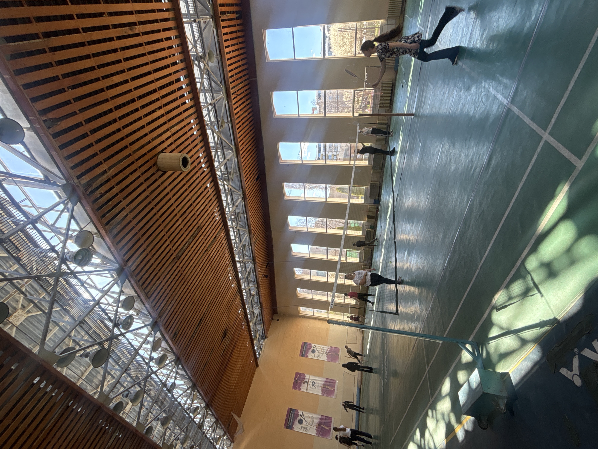A tilted, wide-angle shot inside a gymnasium. The ceiling is a complex structure of wooden slats and metal supports with numerous lighting fixtures. Natural light streams in through a series of tall windows along one wall. A badminton net divides the green court. Several people are present, some appearing to be engaged in a badminton game. Posters are affixed to the wall beneath the windows. The gymnasium likely is a public facility, possibly in a public gym, located in Sofia, Bulgaria, given the language on the posters.