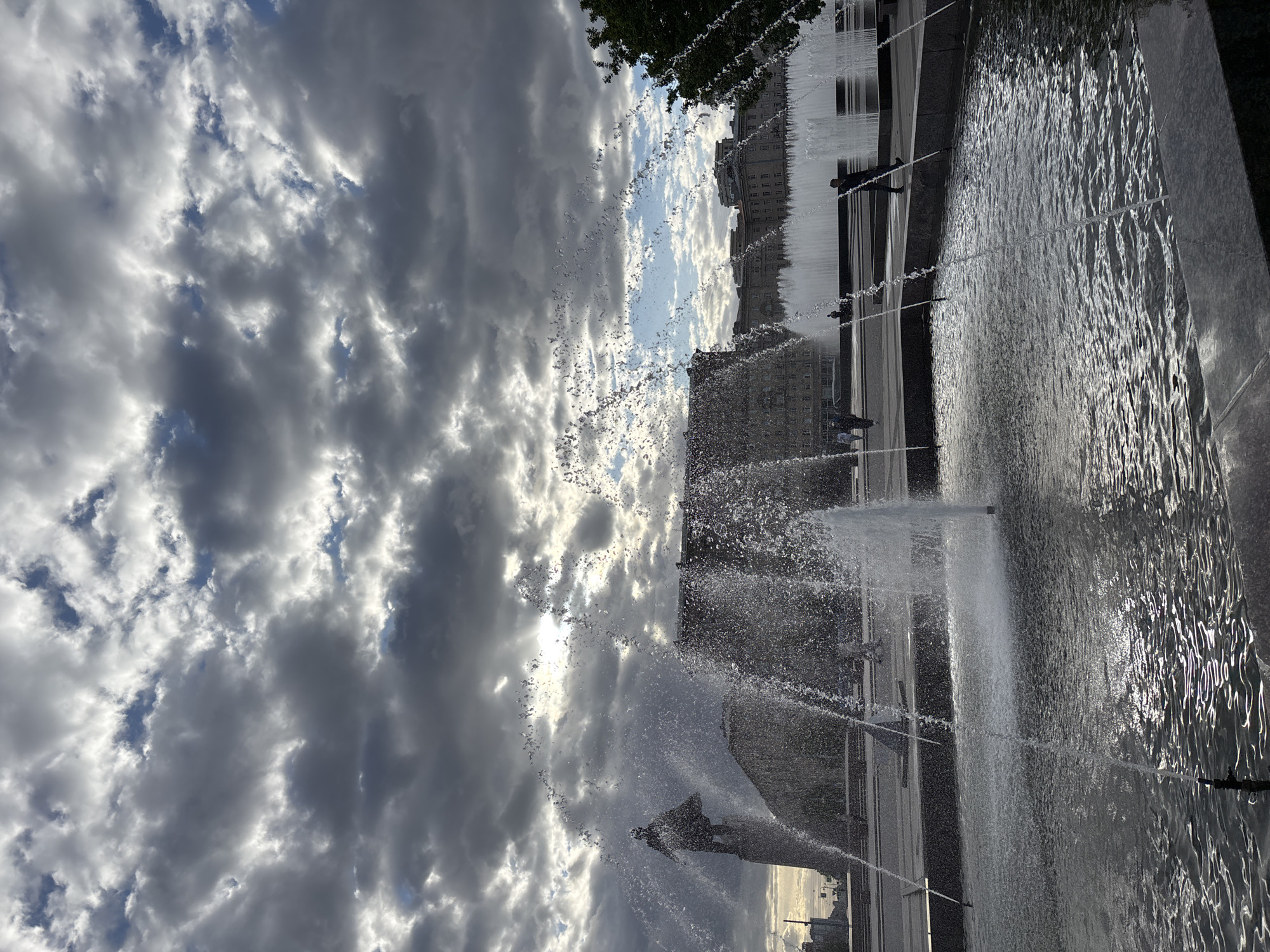 The image captures a fountain in Saint Petersburg, Russia. The sky is filled with dramatic clouds, partially obscuring the sun. Water jets spray upwards from the fountain, creating a mist in the air. In the background, blurred buildings are visible behind the fountain. People can be seen walking along the edge of the fountain basin. The water in the fountain basin reflects light, creating patterns and highlights.