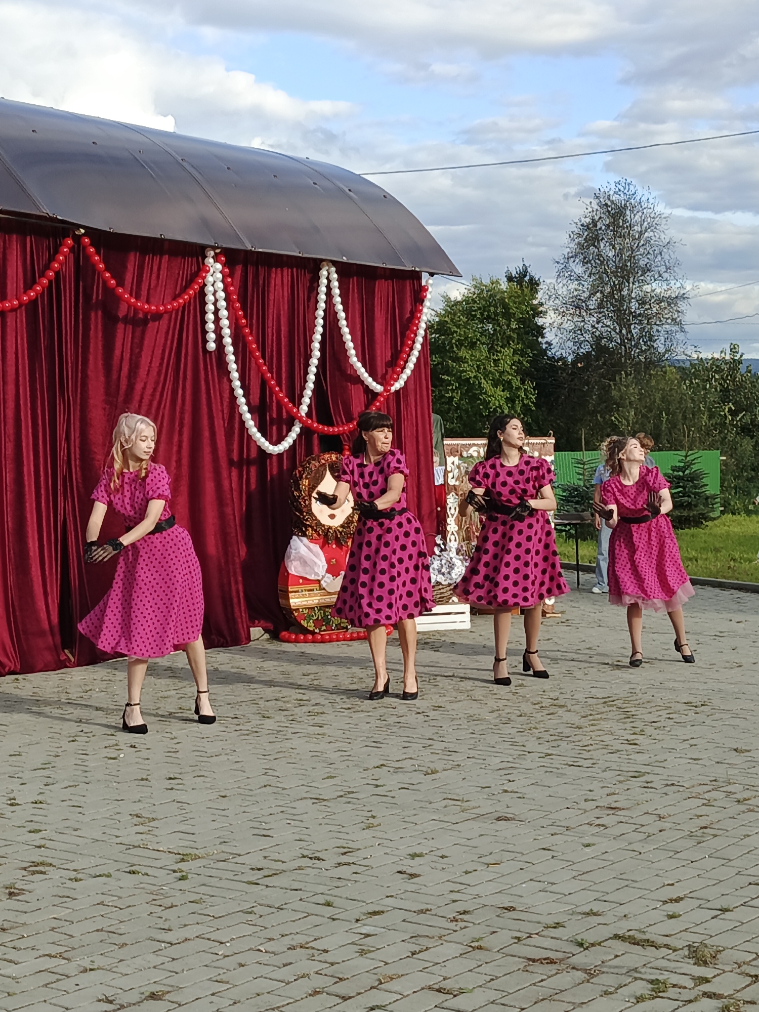 Four women in pink polka-dot dresses are performing on a paved outdoor stage in front of a red curtain backdrop. The woman on the far left has blonde hair and is wearing black gloves. The second woman from the left has dark hair and is also wearing black gloves. The third woman from the left has dark hair and is wearing black gloves. The woman on the far right has light brown hair and is wearing black gloves. Behind the performers, a large cutout of a traditional Russian doll (Matryoshka) is visible. The stage appears to be set up near a tree-lined area with a cloudy sky. The scene is likely taking place in Medvedyovka, Russia.