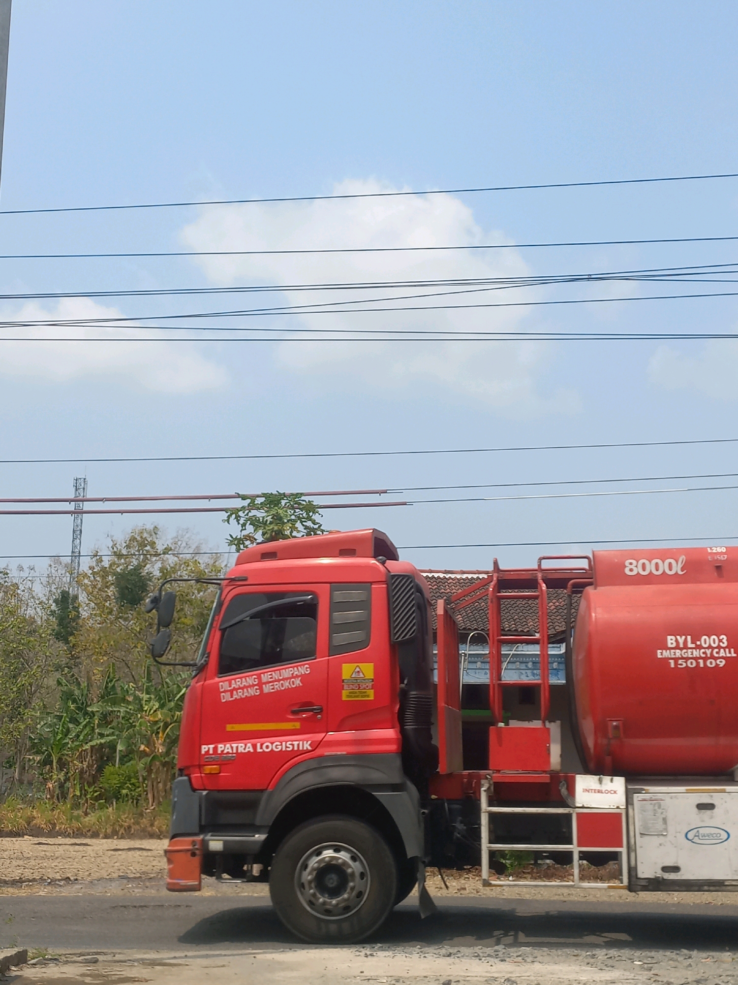 A red tanker truck with "PT PATRA LOGISTIK" written on its side is parked on a dirt road. The truck has a large cylindrical tank on its back. The tank is also red and has markings that read "8000L" and "BYL-003 EMERGENCY CALL 150109". Above the truck, several power lines are strung across a blue sky with some white clouds. In the background, there are trees and a cell tower. The overall scene suggests an industrial or transportation-related setting in Baturetno, Indonesia.