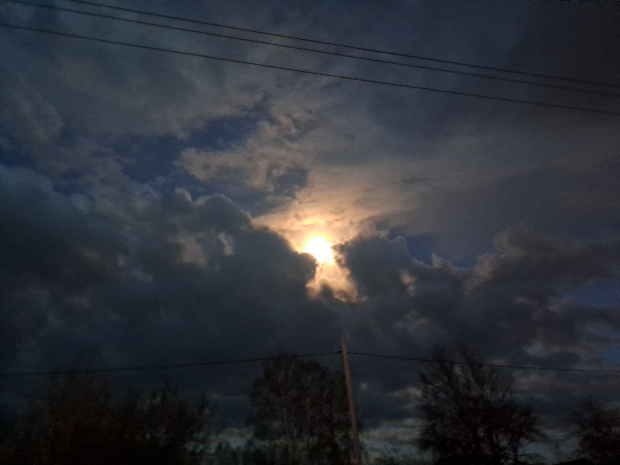 The image displays an atmospheric sky scene, dominated by a dynamic interplay of dark, heavy clouds and brilliant light. A powerful, intense light source, likely the sun, is partially obscured by the dense cloud cover, creating a radiant burst of light that illuminates the surrounding cloud edges with a golden glow. Patches of lighter blue sky are visible in areas where the clouds part.
Several dark, thin lines, resembling power or telephone cables, traverse horizontally across the upper and middle sections of the sky. Towards the lower right, a distinct vertical pole or post extends upwards. The bottom portion of the frame features the dark, silhouetted forms of trees or bushes, indistinct against the muted light from below.
The scene captures a transient moment in the weather, with the dramatic sunbeam piercing through the overcast sky suggesting either an early morning, late afternoon, or a break in a heavily clouded day. The mood evoked is one of natural grandeur and a hint of a changing atmosphere. There is no visible human activity or text in the image. The setting appears to be an outdoor, possibly rural or semi-rural landscape, consistent with the provided location context of Полянки, Russia.