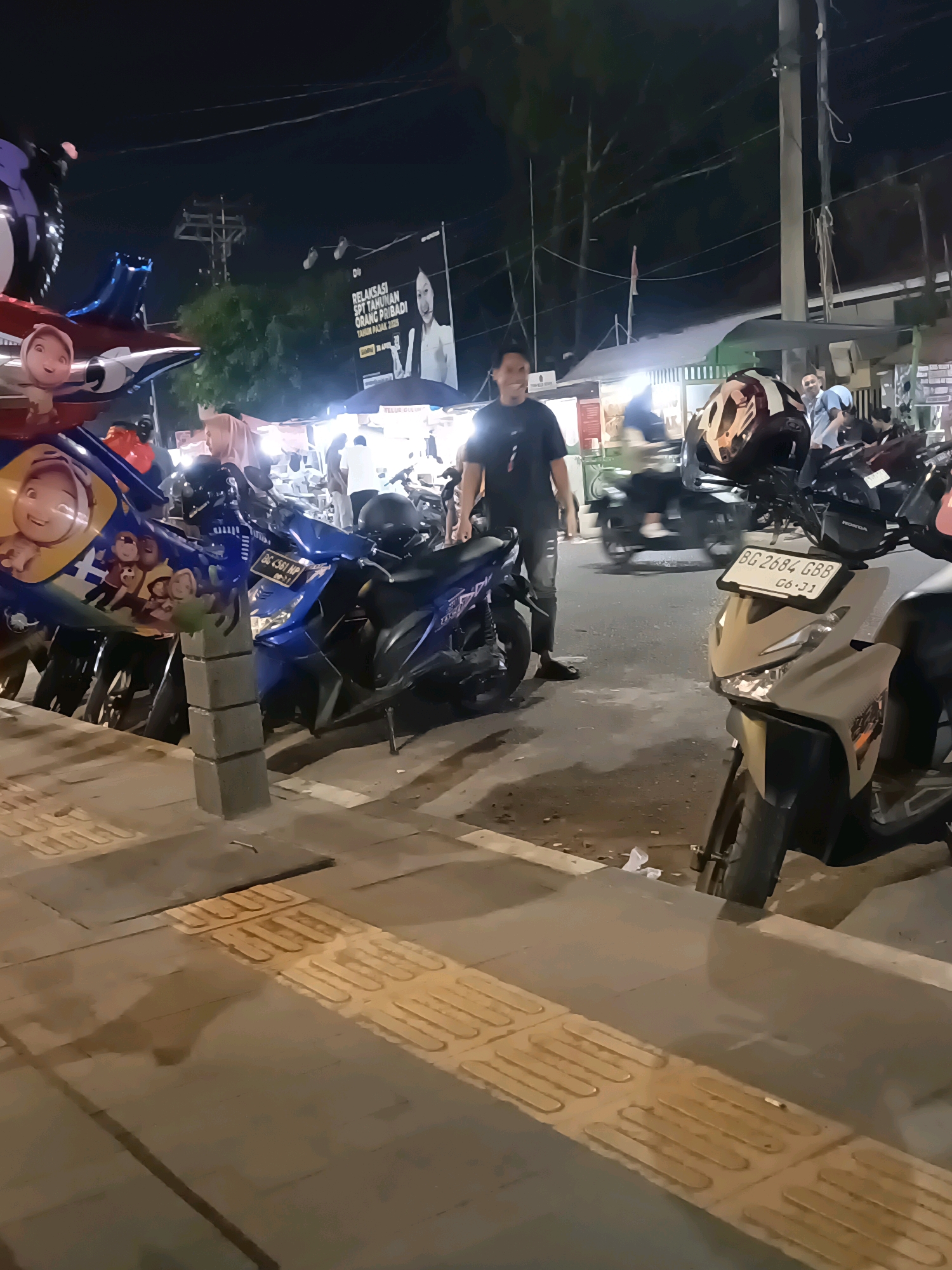 The image depicts a street scene at night in the city of Lubuklinggau, Indonesia, likely in a market or commercial area. In the foreground, a sidewalk with tactile paving is visible, with several motorcycles parked on the street adjacent to it. One light-colored scooter in the right foreground has a license plate reading "BG 2684 GBB" and "06-31". To the left, a blue and yellow decorated motorcycle is also parked. A man in a black t-shirt and dark pants is standing on the sidewalk, looking towards the right. Further back, other individuals are visible, some near stalls or shops. A large banner with text and an image of a woman is displayed in the background, with trees and utility poles visible against the night sky. The lighting suggests artificial sources from the stalls and streetlights.