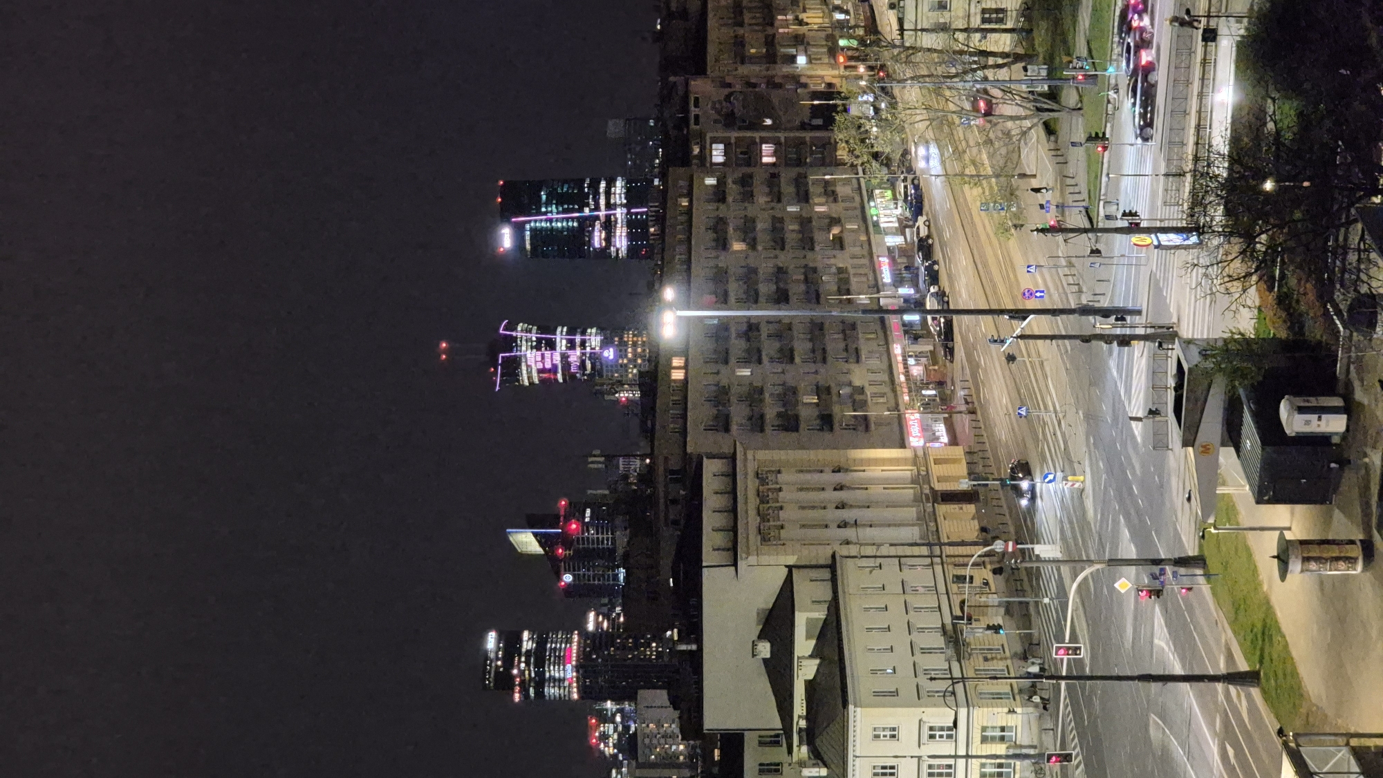 The image captures a night view of Warsaw, Poland. On the left, illuminated skyscrapers rise against the dark sky, their lights reflected in the city below. Adjacent to the skyscrapers are residential buildings. To the right, an empty street stretches into the distance, marked with traffic lights and signs. A metro station is visible on the right side of the street. There is an absence of people.