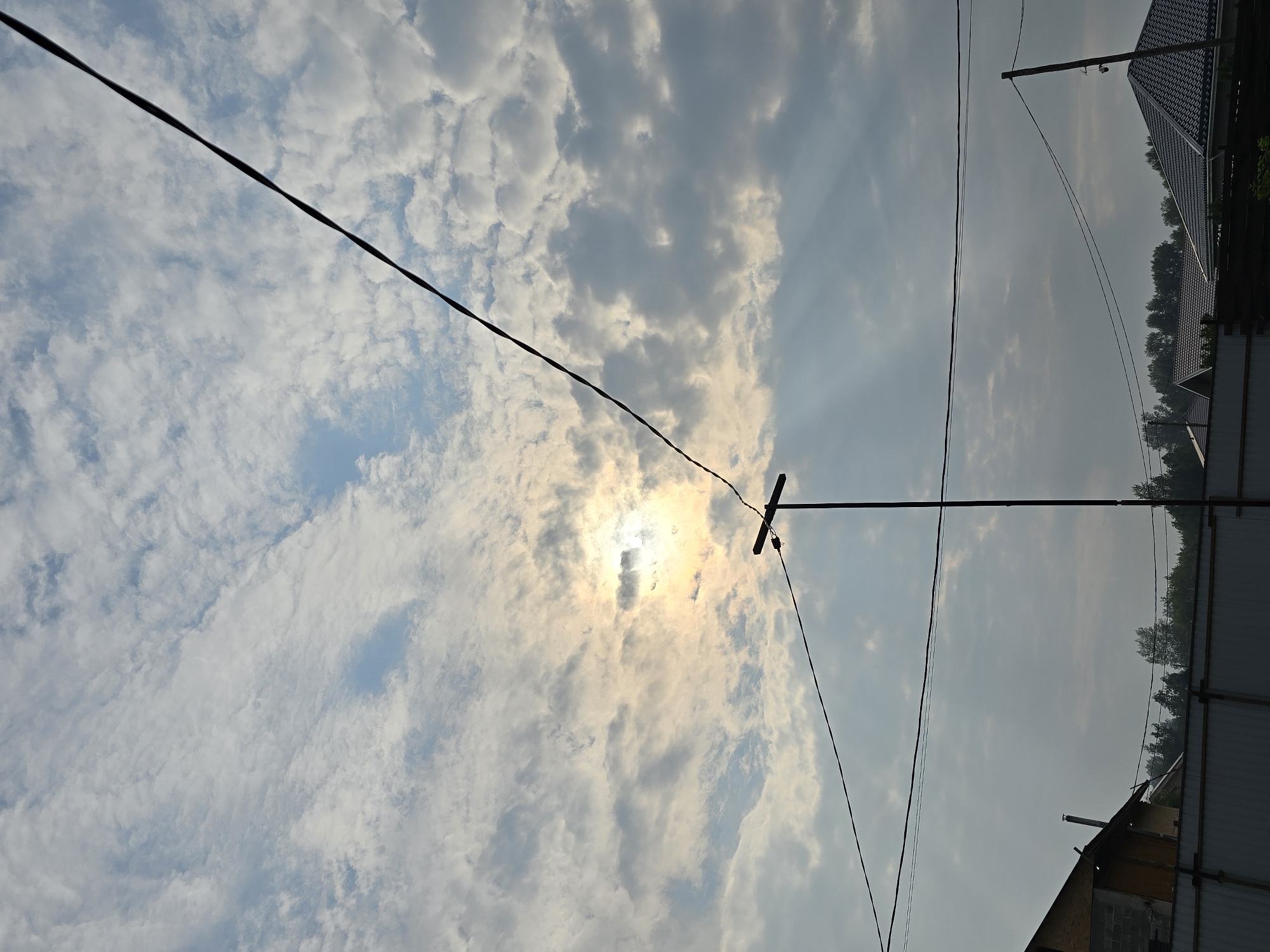The image depicts an outdoor scene under a partially cloudy sky, with the sun visible through a break in the clouds, casting rays of light. In the foreground, there are power lines crossing the frame, supported by poles. A section of a fence is visible at the bottom, with parts of houses and trees behind it. The architecture of the houses appears to be typical of a suburban or rural setting. The lighting suggests it is either early morning or late afternoon. The scene is located in Ермолаево, Russia. There are no people visible in the image.
