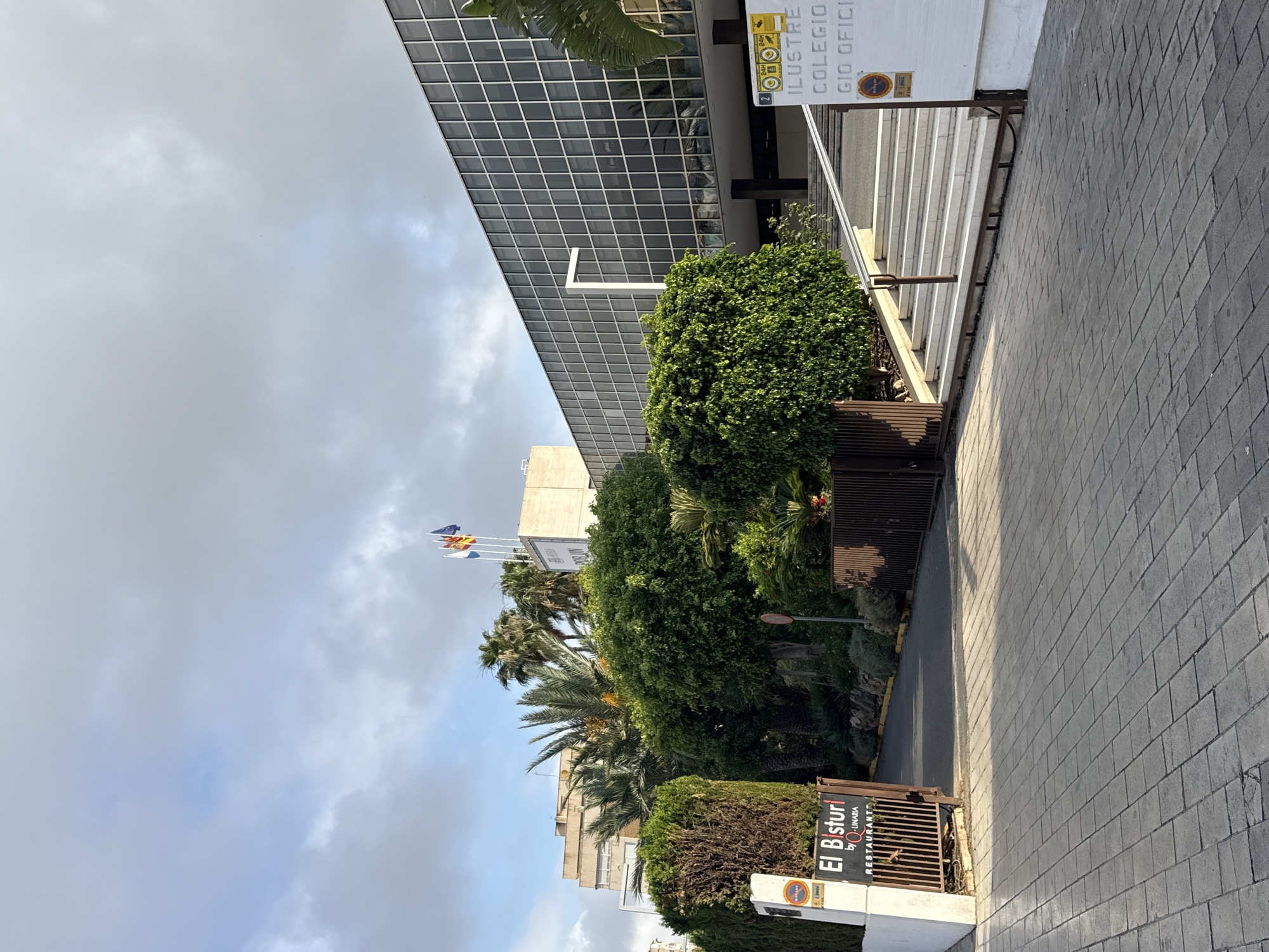 The image shows a modern building with a glass facade and a white accent structure on the left, set against a cloudy sky. In the foreground, there is a paved walkway and a set of stairs leading up to the building. To the left of the stairs is a white pillar with signage, including "El Bisturi by Q-LINARIA RESTAURANT". Beyond the paved area, there is lush green foliage, including trees and bushes, and a partial view of a fence. The building appears to be an institutional or commercial establishment, with flags flying from its roof. Another building, possibly residential, is visible in the background. The scene suggests an urban or suburban environment.