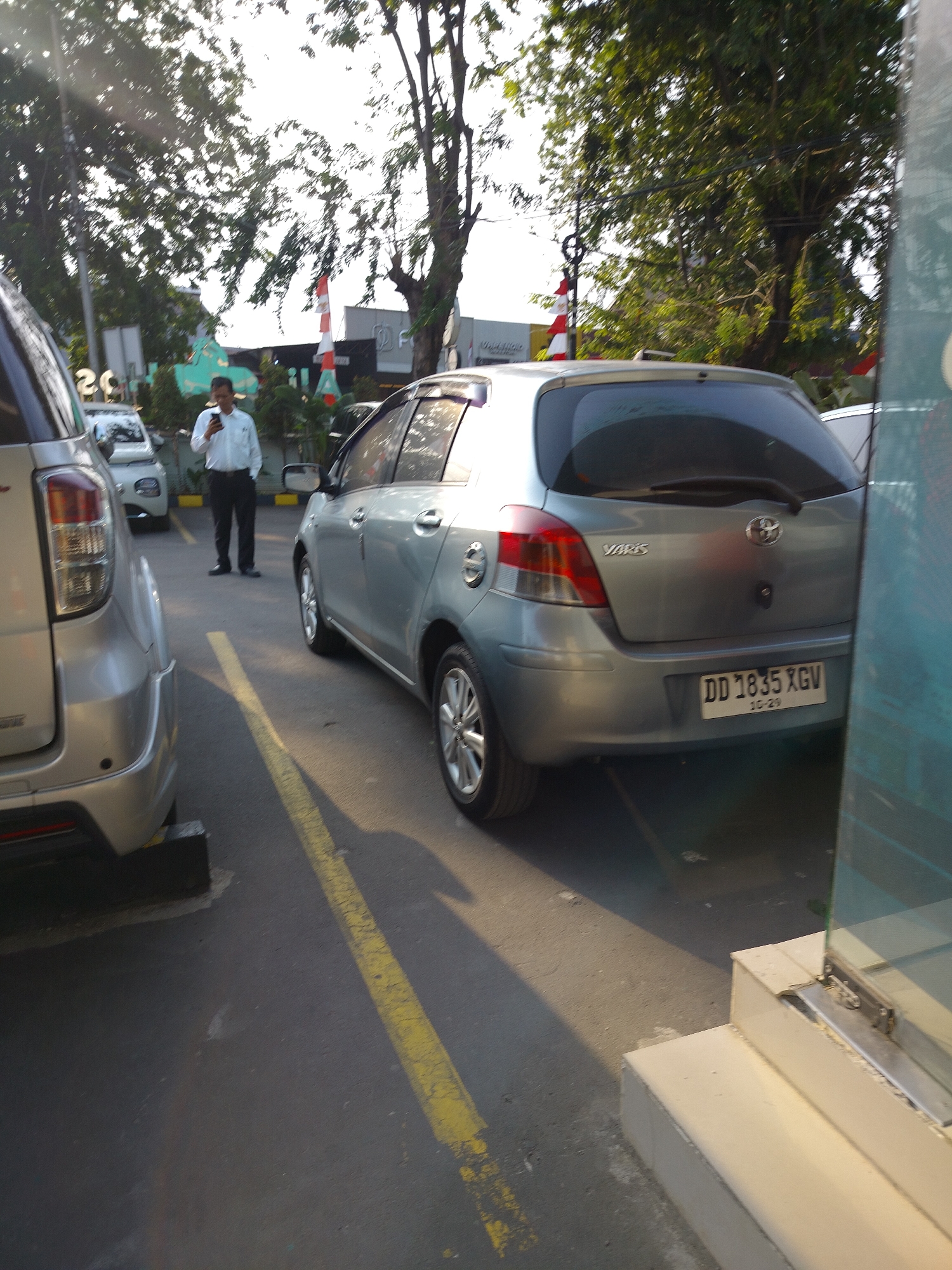 A silver Toyota Yaris, license plate DD 1835 XGV, is parked on an asphalt surface marked with yellow lines. The car's rear is visible, showing its taillights, emblem, and license plate. A man in a white long-sleeved shirt and black trousers stands to the left of the Yaris, holding a mobile phone. He appears to be looking at the device. To the left of the Yaris, another silver vehicle, a different model, is partially visible. The scene is outdoors, with trees and buildings in the background. The lighting suggests it is late afternoon or early evening. The location is identified as East Jakarta, Indonesia.