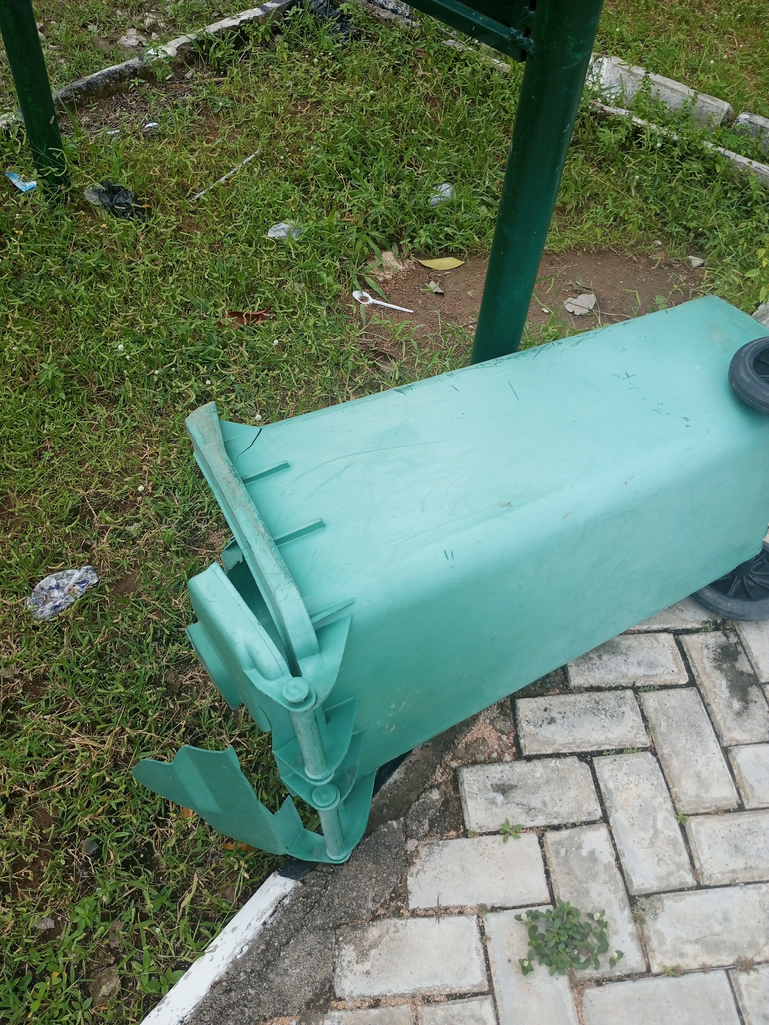 A broken, green plastic garbage bin lies on its side on a paved walkway and adjacent grassy area. The bin is damaged, with a large section of its front broken off and hanging. Two black wheels are visible on the right side of the bin. The walkway is made of light-colored rectangular pavers, and the grass is green and somewhat overgrown. In the background, a green metal pole supports an unseen structure. Scattered litter, including plastic bags and wrappers, is visible on the grass. The scene appears to be in an outdoor public space in Jalingo, Nigeria.