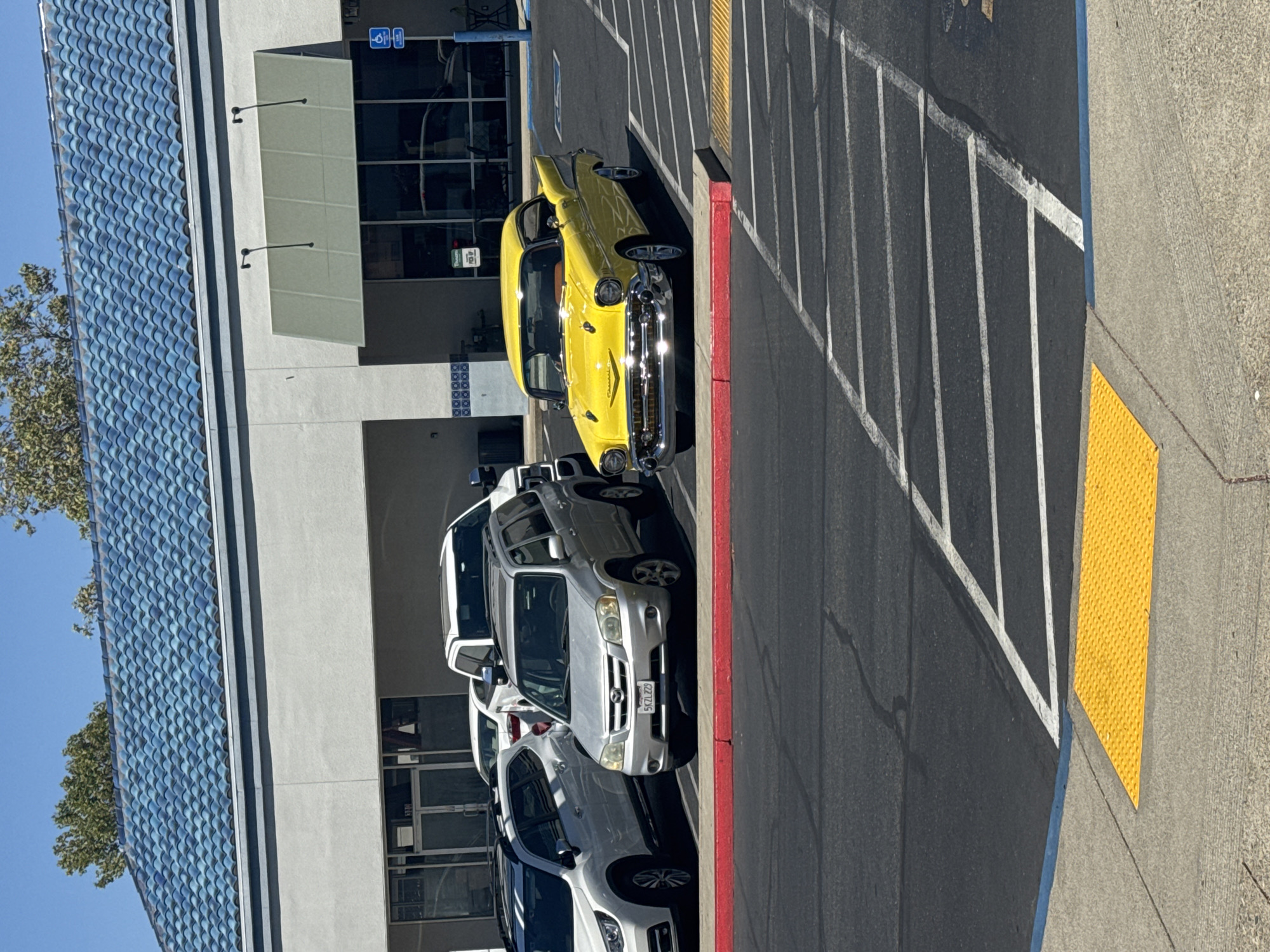 A bright yellow classic car is parked in a parking lot in Roseville, California. The car, a Chevrolet Bel Air, is a two-door coupe with chrome accents and whitewall tires. It is parked next to a silver SUV and a white SUV. The building in the background has a blue tiled roof and a light-colored facade. The parking lot is asphalt with white painted lines and a yellow tactile paving surface at the edge of a sidewalk. The sky is clear and blue, and the sun is shining, casting shadows. There are no people visible in the image.
