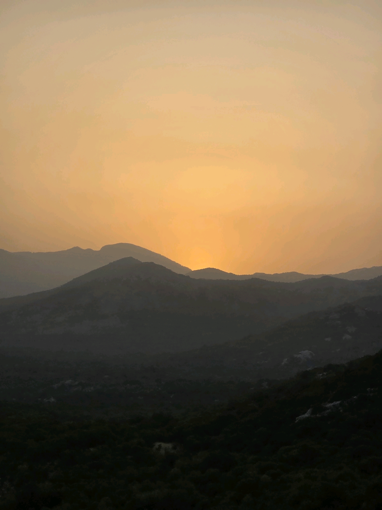 The image depicts a serene mountain landscape at sunset, likely taken in the vicinity of Medun, Montenegro. The sky is a warm gradient of orange and yellow, with the sun partially obscured by the silhouetted mountain ranges in the foreground. Multiple layers of mountains recede into the distance, their peaks softly defined against the luminous sky. The lower slopes are covered in dark, dense vegetation, creating a stark contrast with the bright sky. There are no visible people, animals, or man-made structures in this scene, emphasizing the natural beauty and tranquility of the mountainous environment. The overall impression is one of peaceful solitude and the grandeur of nature at dusk.