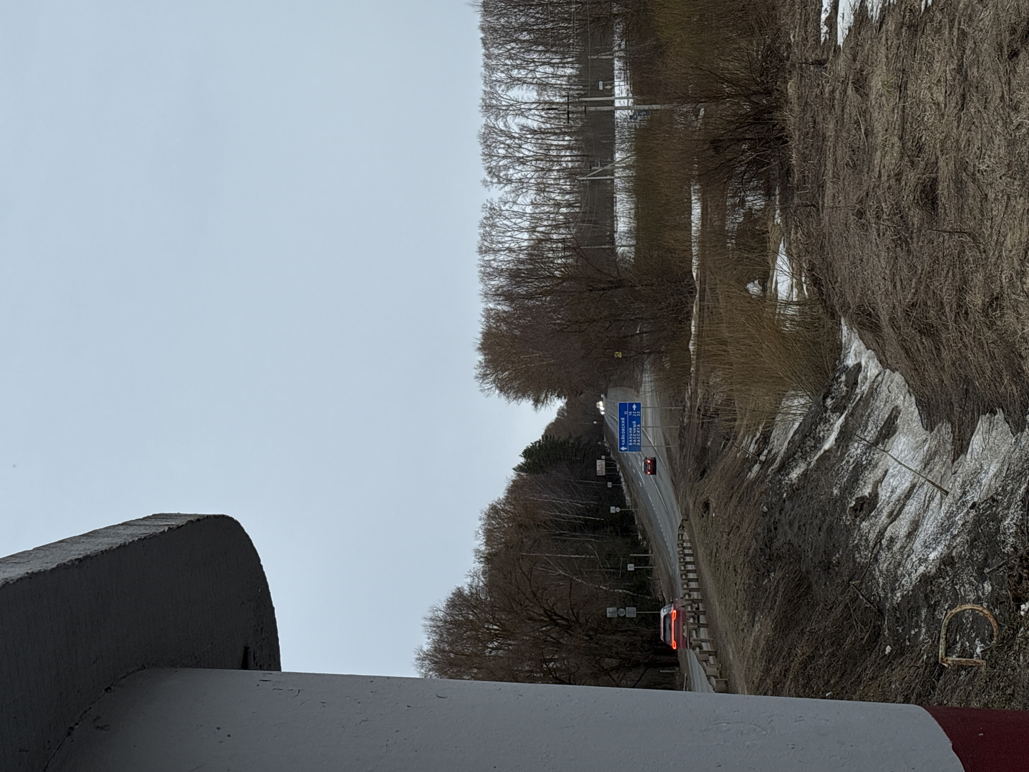 The image shows a view of a road from an elevated position, likely a bridge or overpass, in what appears to be a rural or semi-rural area. The road has two lanes, with signage visible indicating directions to destinations like "Chaykovskiy," "Zarechnyy," and "Vesnota," with distances in kilometers. The signage and names suggest the location is near or related to the city of Rusalevka, Russia. A red car is stopped on the road, possibly at a traffic light. The surrounding environment includes trees and vegetation, with some patches of snow on the ground. Part of a concrete structure is visible in the foreground. The sky is overcast.