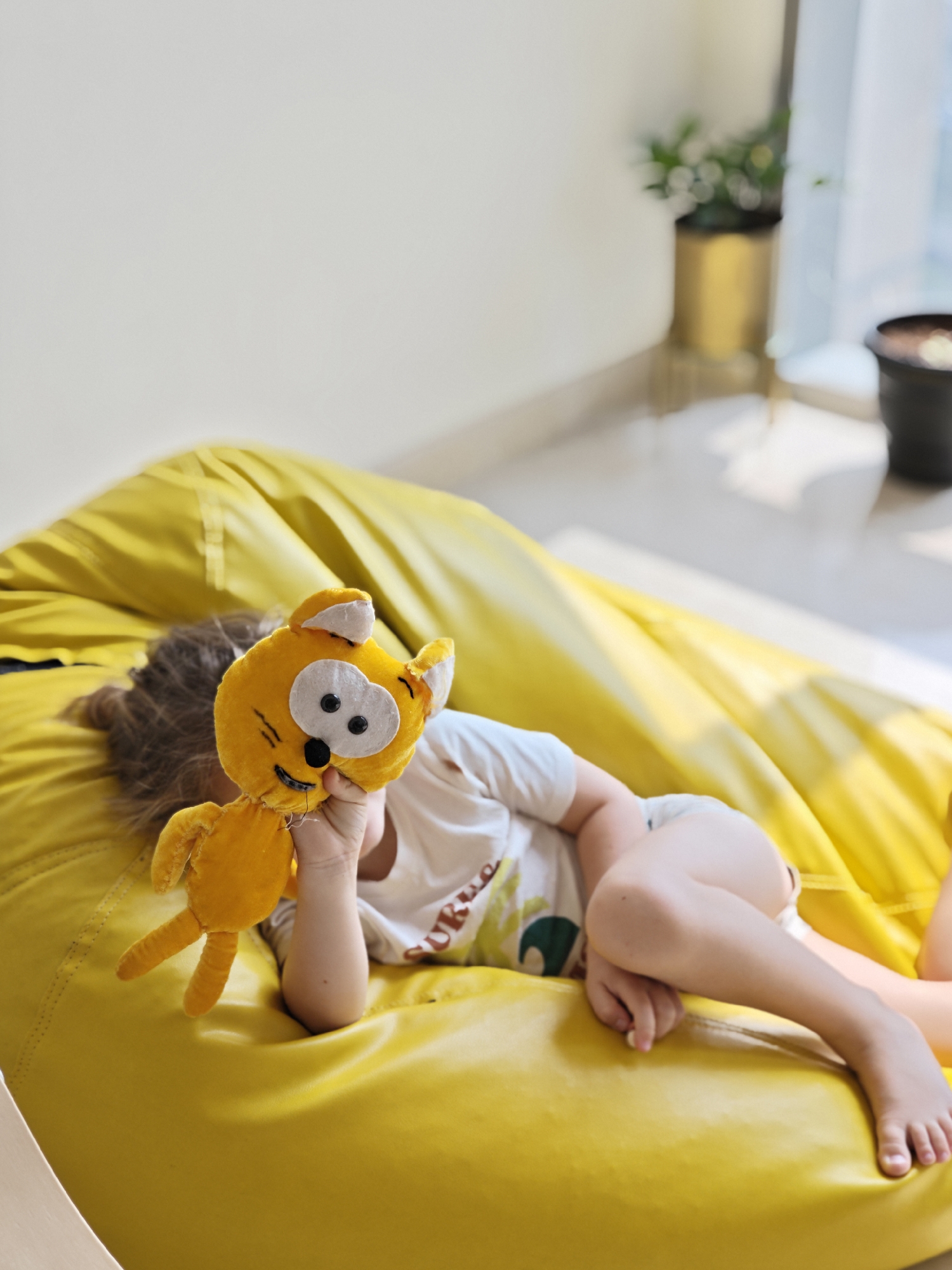 A child lies on a yellow beanbag chair, holding a yellow plush cat toy. The location appears to be indoors, with a white wall and a window in the background, suggesting a domestic setting. Two potted plants stand near the window. The child is wearing a white t-shirt. The room is filled with natural light. There is no indication of it being in Mumbai, India.