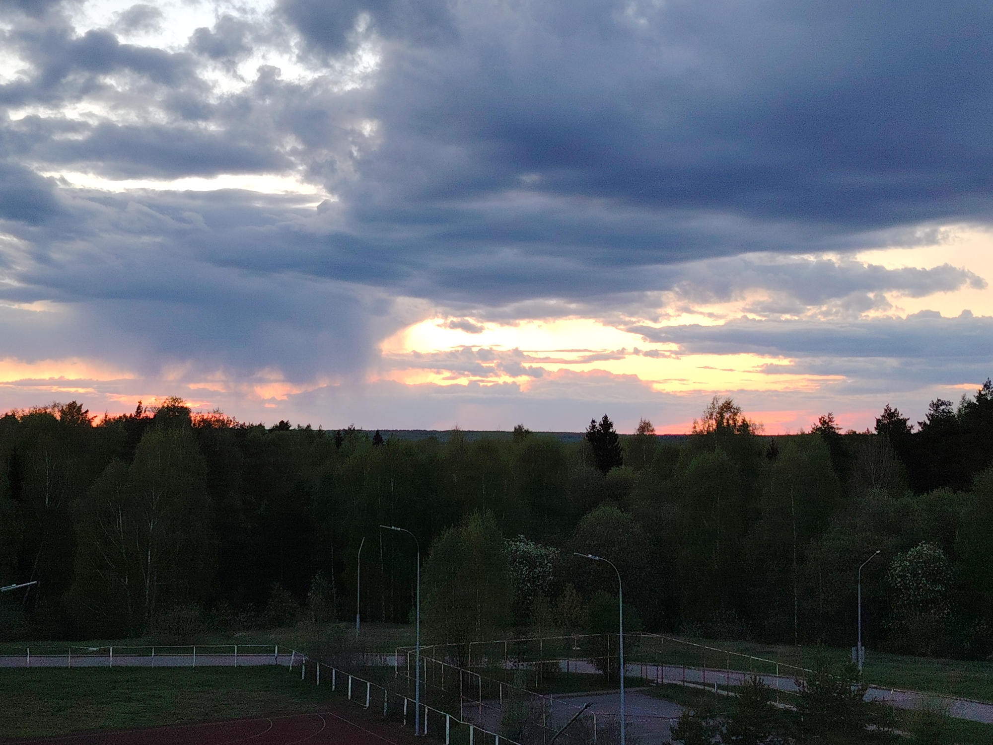The image shows a view of a forest and the sky during a sunset or sunrise. The sky is filled with dramatic clouds, with a mix of gray, dark blue, and orange hues from the setting or rising sun. The forest is dense with mostly green trees. In the foreground, there appears to be a sports field or track with metal fencing and some street lights.
The provided location, Марковский, Russia, is relevant to the overall environment.