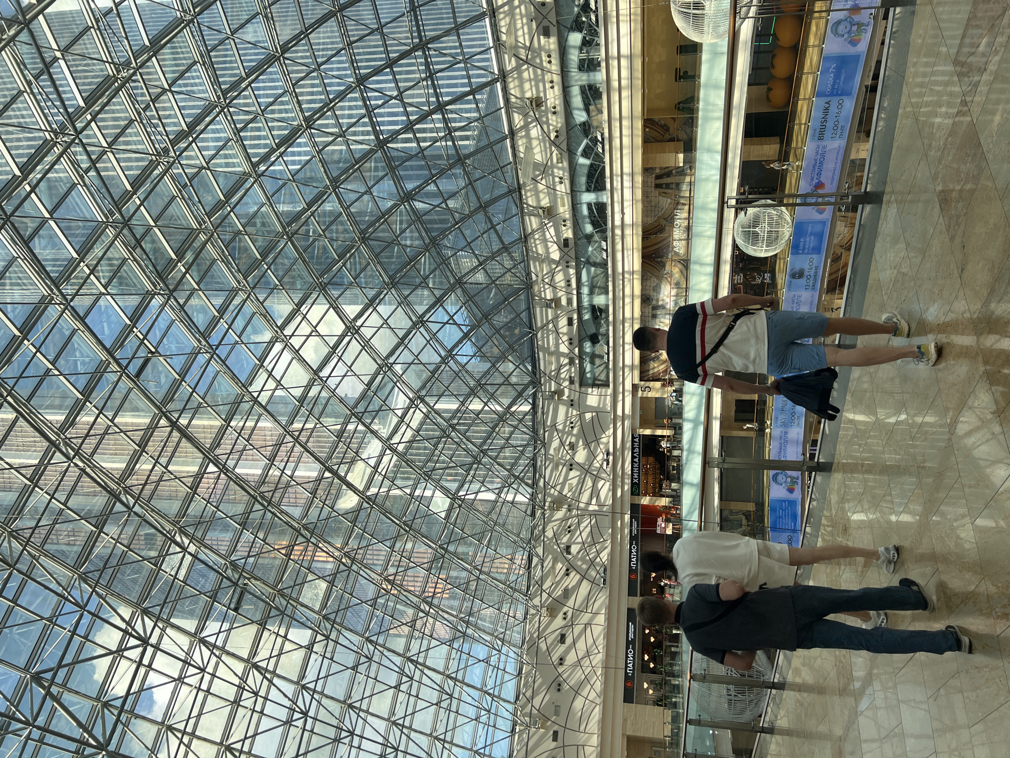 Three individuals are walking on a polished floor within a modern shopping mall. The mall features a large, domed glass and metal roof structure, reflecting the sky and surrounding skyscrapers. One person, wearing a dark t-shirt and jeans, walks away from the camera. Another, in a white t-shirt and shorts, walks ahead of the first person. The third person, in a striped t-shirt and blue shorts, walks slightly behind and to the right of the other two, carrying a dark bag. Multiple levels of the mall are visible, with shops and signage, including "ПАТИО" and "ХИНКАЛЬНАЯ," indicating food establishments. Decorative spherical light fixtures are suspended from the railings. The architecture suggests a contemporary urban environment, consistent with the city of Khimki, Russia.