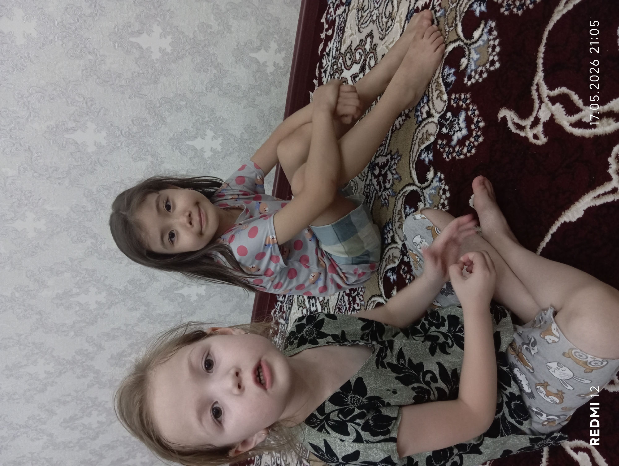Two young girls are sitting on a patterned rug in what appears to be an indoor setting, possibly a home. The girl in the foreground has blonde hair and is wearing a dark floral-patterned top with animal-print shorts. The girl in the background has dark hair and is wearing a light-colored top with red polka dots and plaid shorts. The background features a wallpapered wall. The photograph seems to have been taken in Bekabad, Uzbekistan, based on the metadata in the image. The date and time of the photograph are 17.05.2026 21:05.