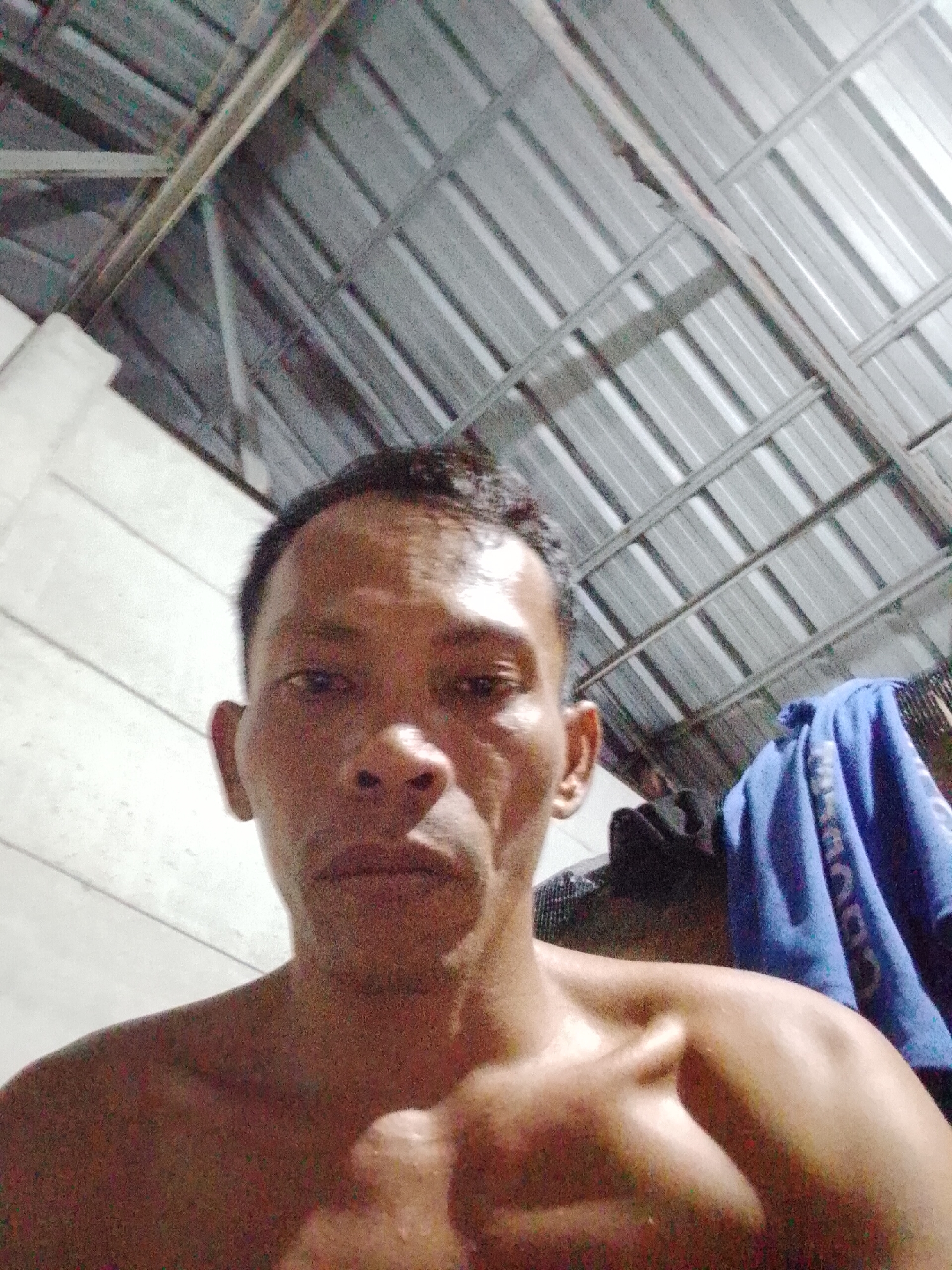 The image shows a man with short, dark hair in a room with a corrugated metal roof and white walls. A blue cloth hangs behind him. The location is in Bukit Ketok, Indonesia.