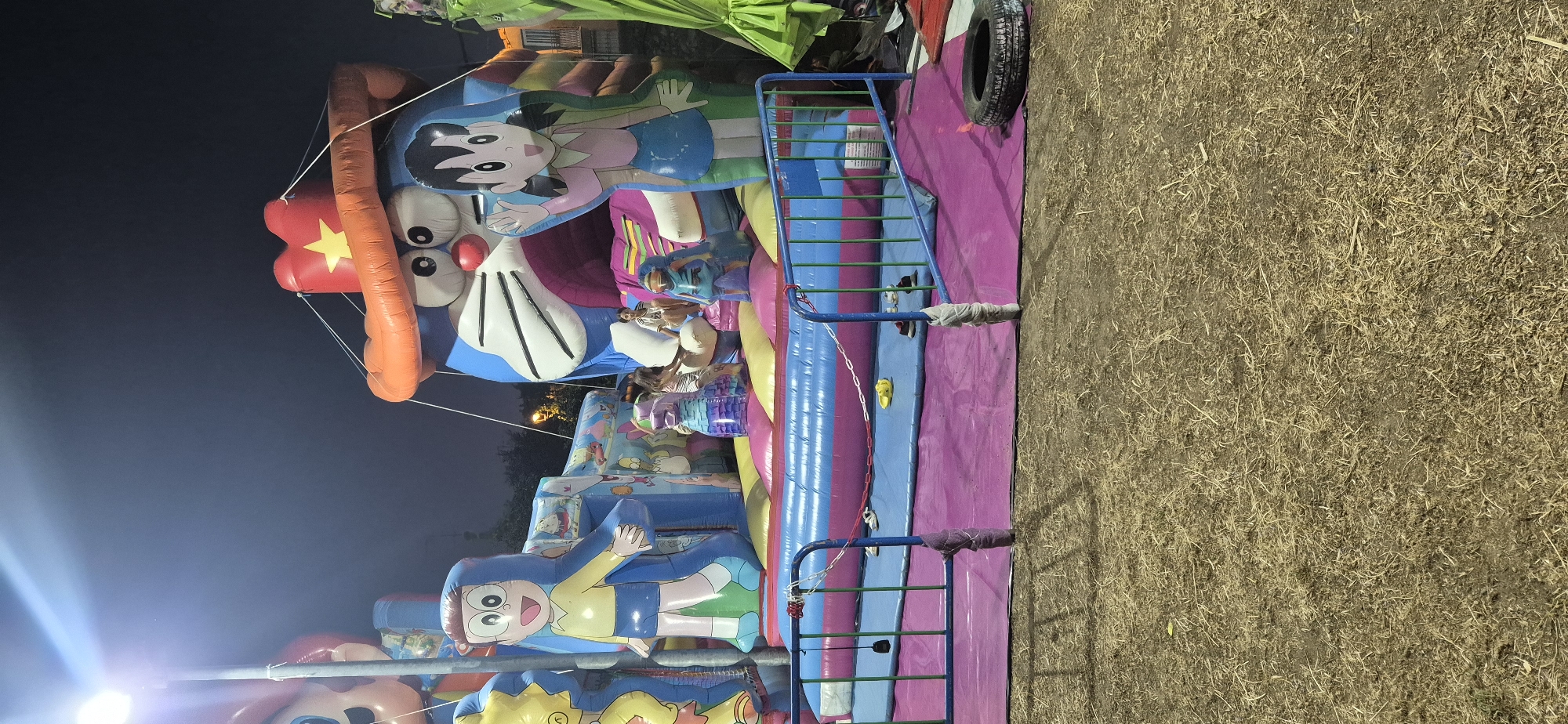 The image depicts an outdoor inflatable playground, likely a bouncy castle, at night. The playground features several large inflatable characters, including a prominent blue character resembling Doraemon wearing a cowboy hat, and other characters that appear to be from popular cartoons. There are children playing on the bouncy castle. The playground is illuminated by artificial lights, creating a bright and festive atmosphere. In the foreground, there is a metallic barrier with a chain and some discarded items like shoes and a tire. The ground around the playground appears to be covered in dry grass or straw. The scene is set in Navaluenga, Spain.
