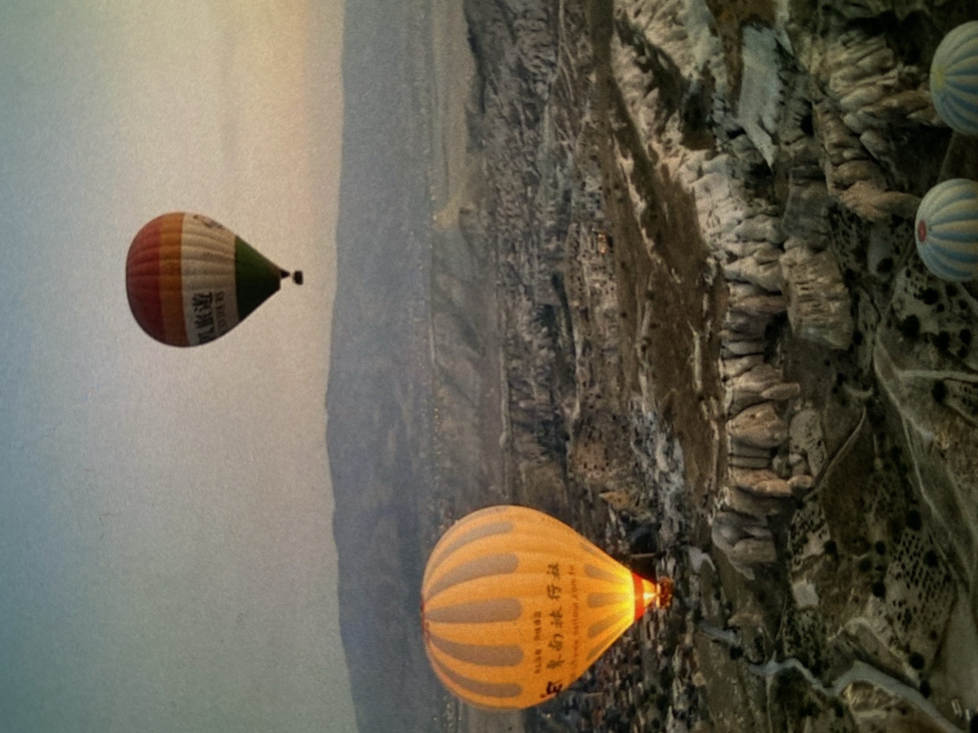 The image, appearing to be rotated 90 degrees clockwise from its original capture, depicts a panoramic view of multiple hot air balloons ascending over a unique, rugged landscape, likely at dawn or dusk due to the soft, diffused lighting and hazy sky.
In the foreground and mid-ground, two prominent hot air balloons are visible. On the left, a balloon with vertical stripes of reddish-brown, white, and green features Chinese characters "浪漫旅程" (Làngmàn lǚchéng), meaning "Romantic Journey," printed on its white section. On the lower right, a large yellow-orange balloon with darker vertical stripes also displays Chinese characters "浪漫之旅" (Làngmàn zhī lǚ), which translates to "Romantic Journey" or "Journey of Romance," along with barely legible English text that appears to include "Cappadocia balloon tour" and a ".tr" internet domain. A bright orange flame from its burner illuminates the lower portion of this balloon. In the upper right background, two smaller, light blue and white striped balloons are partially visible against the rocky terrain.
The setting is characterized by an extensive, arid landscape of distinctive geological formations, commonly known as "fairy chimneys" or eroded rock pillars and valleys. These formations suggest a highly specific location known for such natural wonders. There are hints of paths or roads winding through the terrain and possible cave dwellings or structures carved into the rock formations. The sky in the background is light blue-grey and hazy.
The visible Chinese text "Romantic Journey," the English phrase "Cappadocia balloon tour," and the ".tr" internet domain (indicating Turkey) on the balloons, combined with the unique geological features, strongly suggest the location is Cappadocia, Turkey. This contradicts the provided location context of Bankya, Bulgaria. The event is a popular tourist activity involving hot air balloon rides over this scenic region.