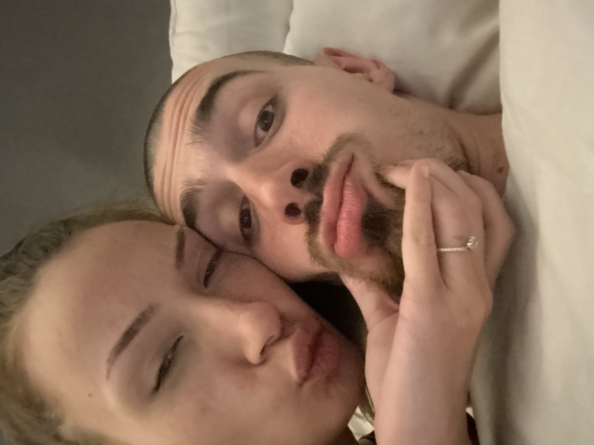 The image displays a close-up view of two individuals, a female and a male, in an intimate indoor setting. The female is positioned on the left, partially cropped, with fair skin and light-colored hair, partially visible at the top. Her eyes are mostly closed, and her lips are pursed into a prominent pout. Her left hand, wearing a thin, silver-colored ring with a small, light-colored stone on her ring finger, is gently cupping the male's chin. The male is on the right, exhibiting fair skin, closely shaved dark hair, a mustache, and a goatee. His right eye is open and directed towards the viewer, while his left eye is partially closed. His lips are also pursed in a pouting expression, mirroring the female's. Both individuals are positioned closely together, suggesting a shared selfie. They are resting on or against white bedding, visible primarily on the right side of the frame and beneath the male. A dark, monochromatic background is visible behind their heads. The image provides no visual information to determine its location in the city of Varna, Bulgaria, or any other specific geographical area.