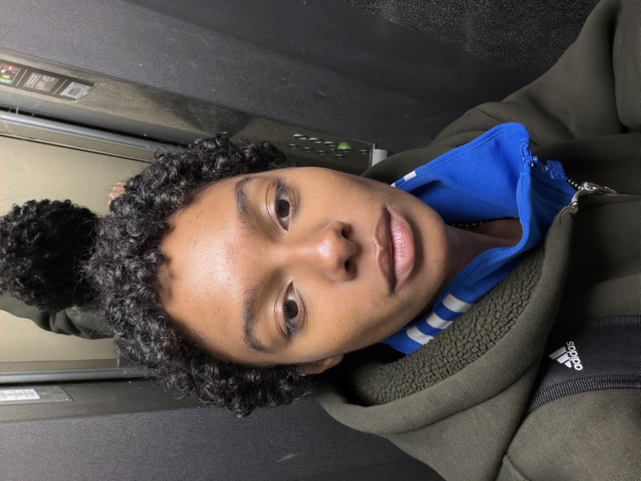 A headshot captures an individual with dark skin and short, tightly curled black hair, looking directly into the camera. The person is wearing a dark olive green, possibly fleece-lined, hooded jacket or sweatshirt. A blue collar with white stripes, partially zipped, is visible beneath the jacket, and a white Adidas logo is present on a black strap over the left shoulder. The individual maintains a neutral facial expression. The scene is set inside an enclosed space, identified as an elevator, with dark, possibly metallic or composite, walls. A control panel featuring various buttons, some illuminated green, and informational stickers is visible on the right side. A partial reflection of the individual's head and upper back is visible in a mirrored surface behind them. This scene occurs within an elevator in the city of Paris, France.