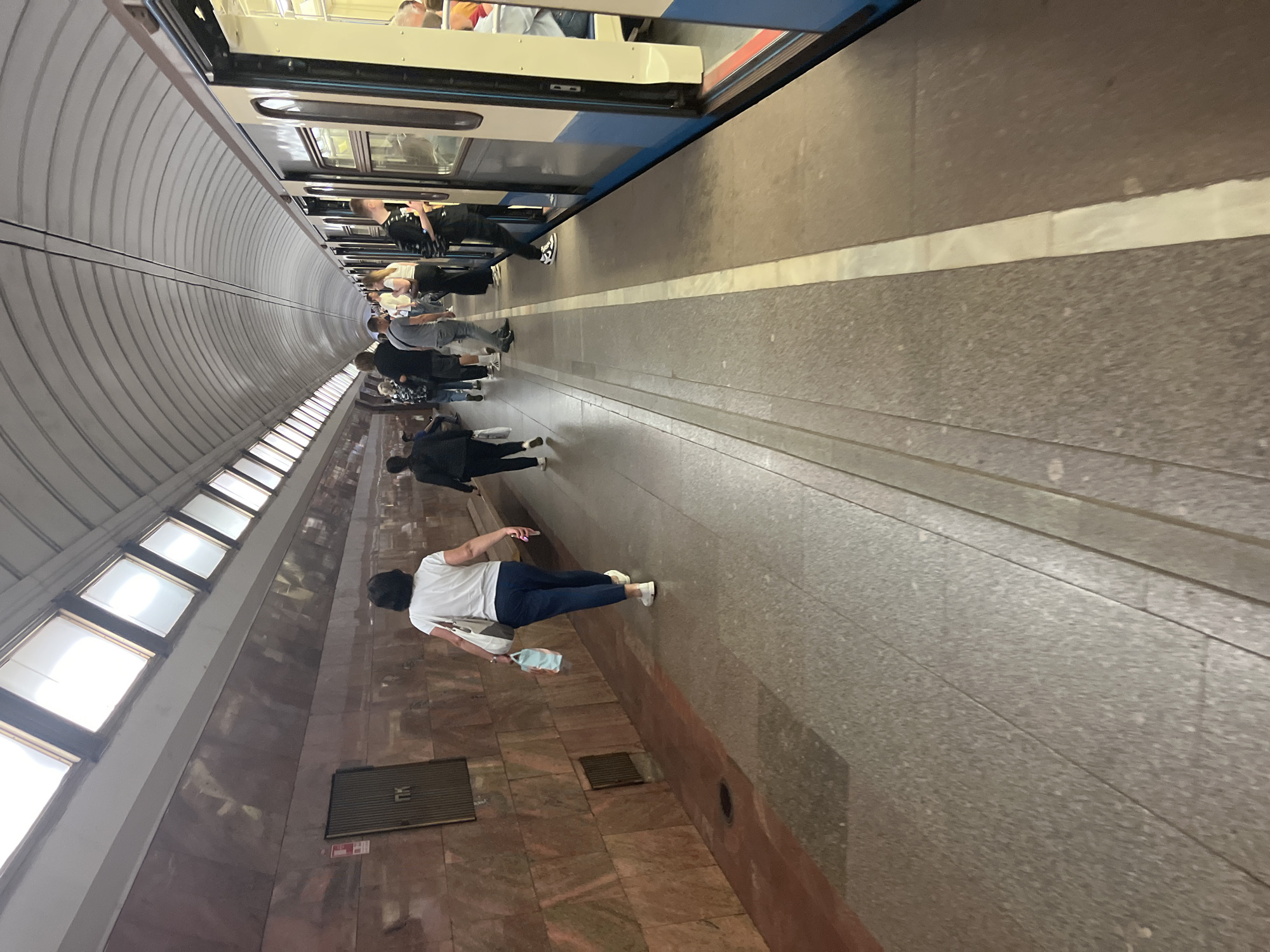 The image depicts a busy subway platform in Moscow, Russia. A train is stopped at the platform, with its doors open and several people boarding and alighting. The platform is made of granite tiles, and the walls are adorned with reddish-brown marble. Overhead, a curved ceiling with a series of windows allows natural light to filter in. On the platform, a woman in a white t-shirt and blue trousers walks away from the camera, carrying a shopping bag. Other passengers are seen standing and walking, some are waiting to board the train, while others are disembarking. The overall atmosphere is one of movement and activity, characteristic of a typical rush hour in a bustling metropolis.