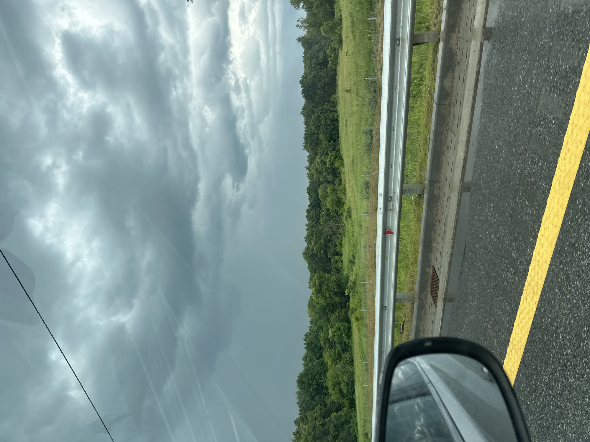 The image depicts a view from a vehicle traveling along a wet, grey asphalt road marked with a bright yellow line. The road is bordered by a metal guardrail and a grassy verge. Beyond the verge, a dense line of lush green trees forms a thick forest. The sky above is overcast with heavy, dark grey clouds, suggesting impending or ongoing rain. The overall scene conveys a sense of a somber, possibly stormy, day. The image appears to be taken while the vehicle is in motion, as indicated by the slight blur and the reflection in the side-view mirror. No people are visible in the frame, and no text is discernable. The setting is rural, with the forest and open grassy areas suggesting a location outside of a city. The weather conditions are clearly the most prominent feature, with the dramatic cloud cover dominating the upper portion of the image and the wet road confirming recent or current precipitation.